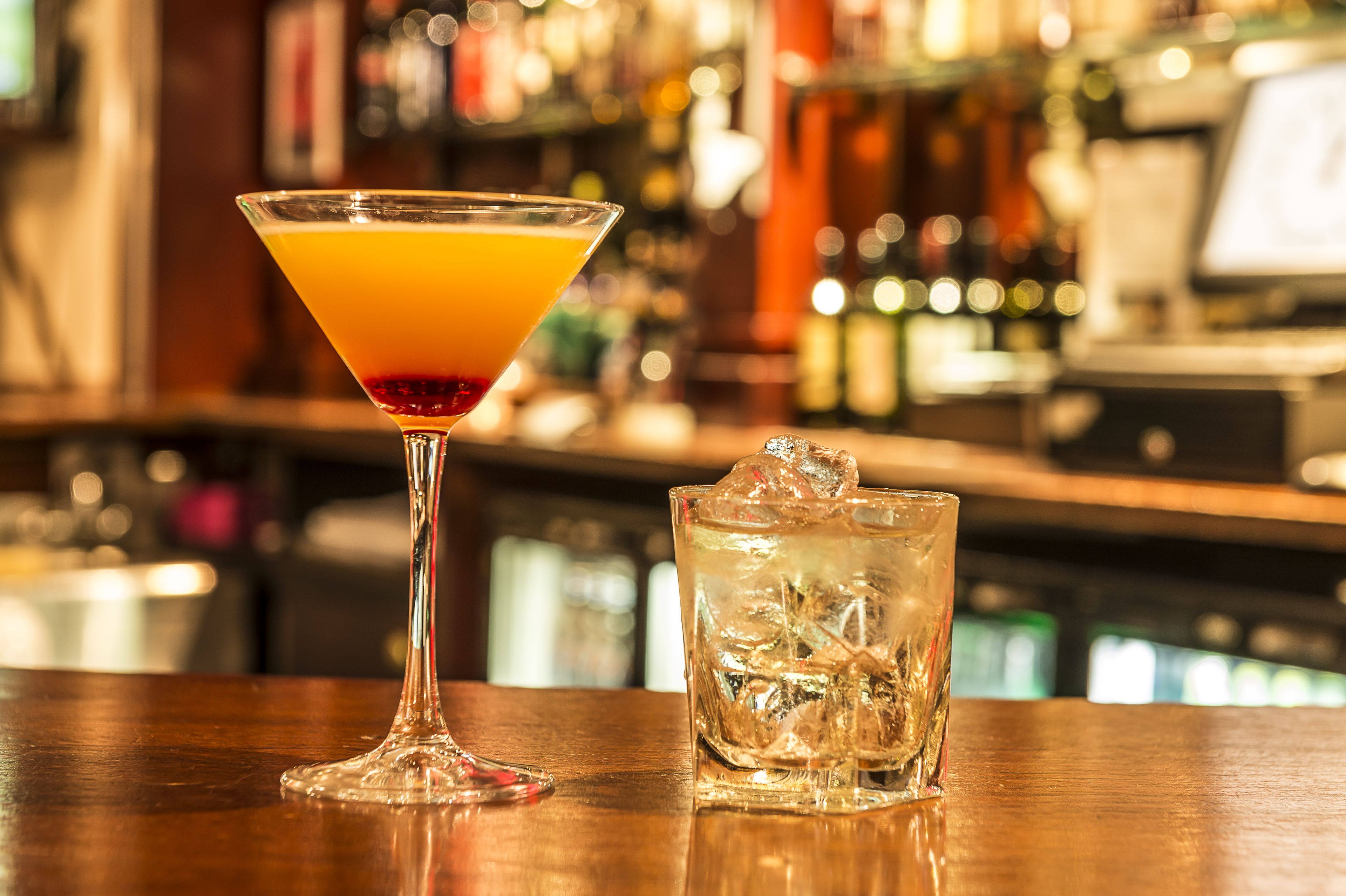 A martini glass and a highball glass with ice cubes on a wooden bar counter.