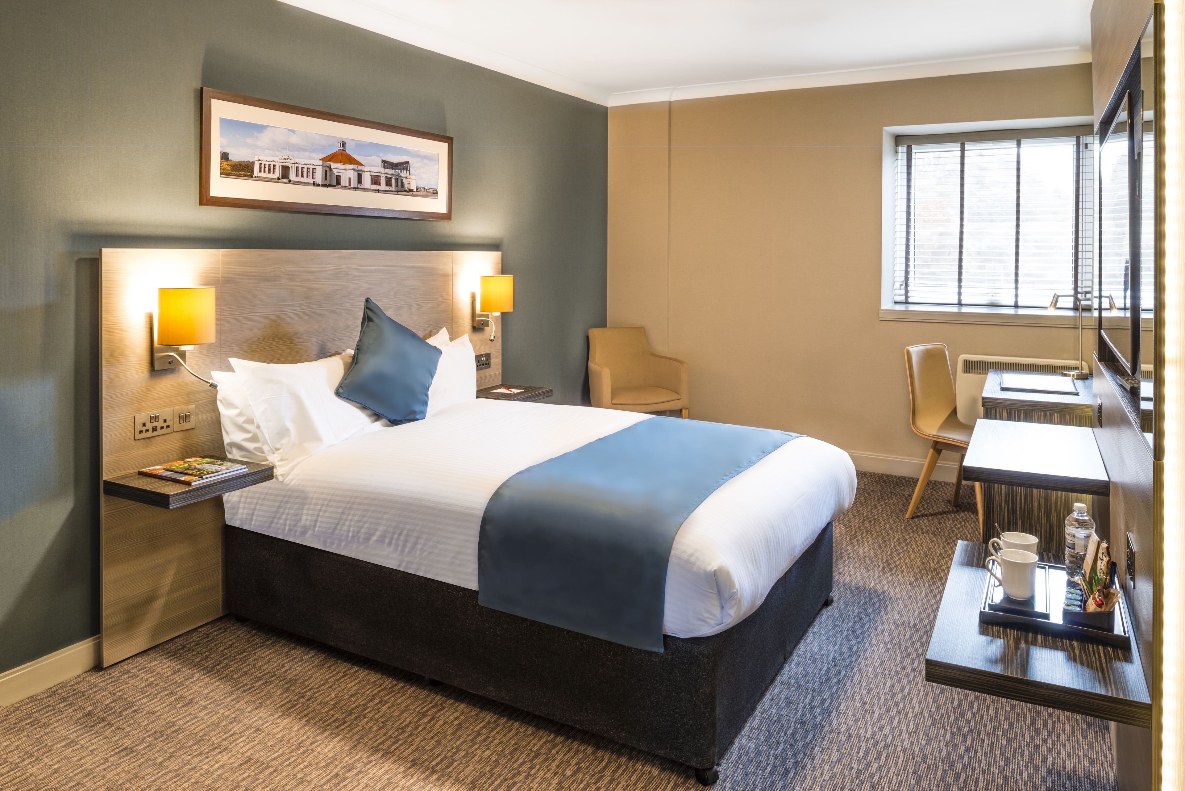Quarto de hotel com uma cama grande, duas luminárias de cabeceira, uma escrivaninha com cadeira, televisão e uma janela com persianas.