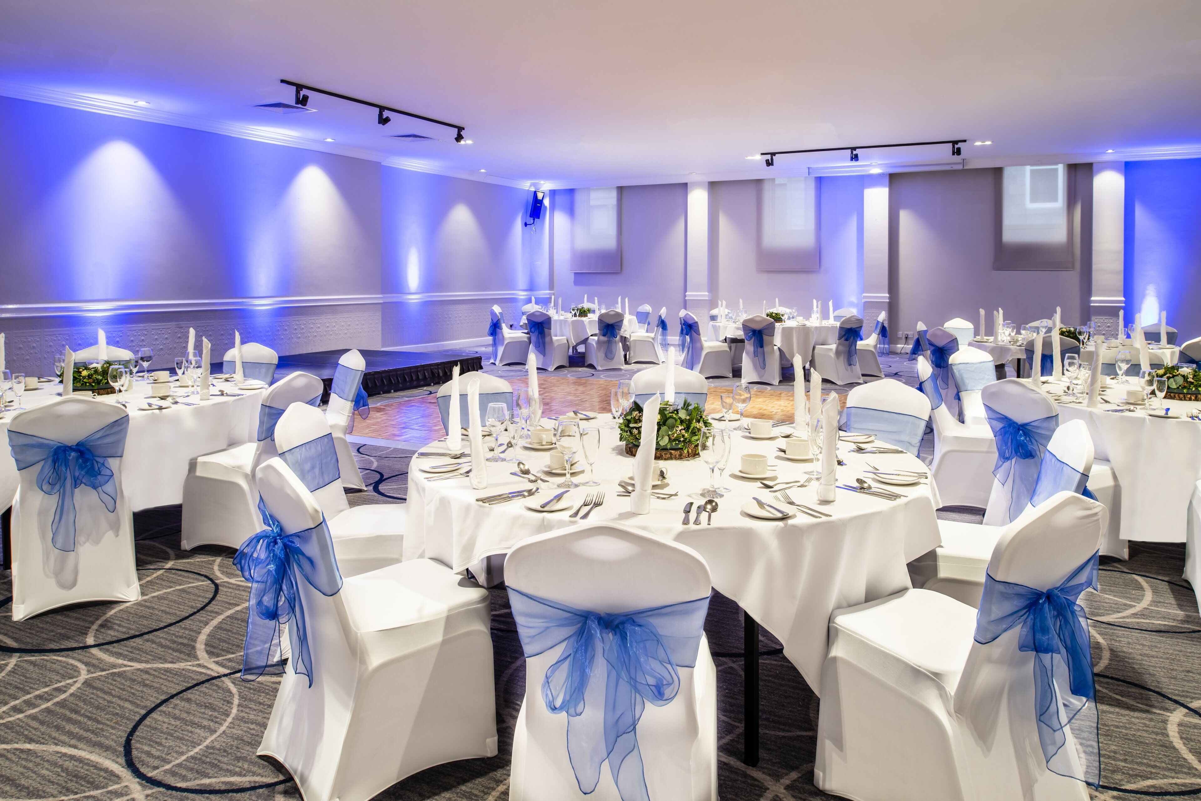 Un salón de banquetes con mesas redondas dispuestas para un evento formal, sillas blancas con cintas azules y un escenario al fondo.