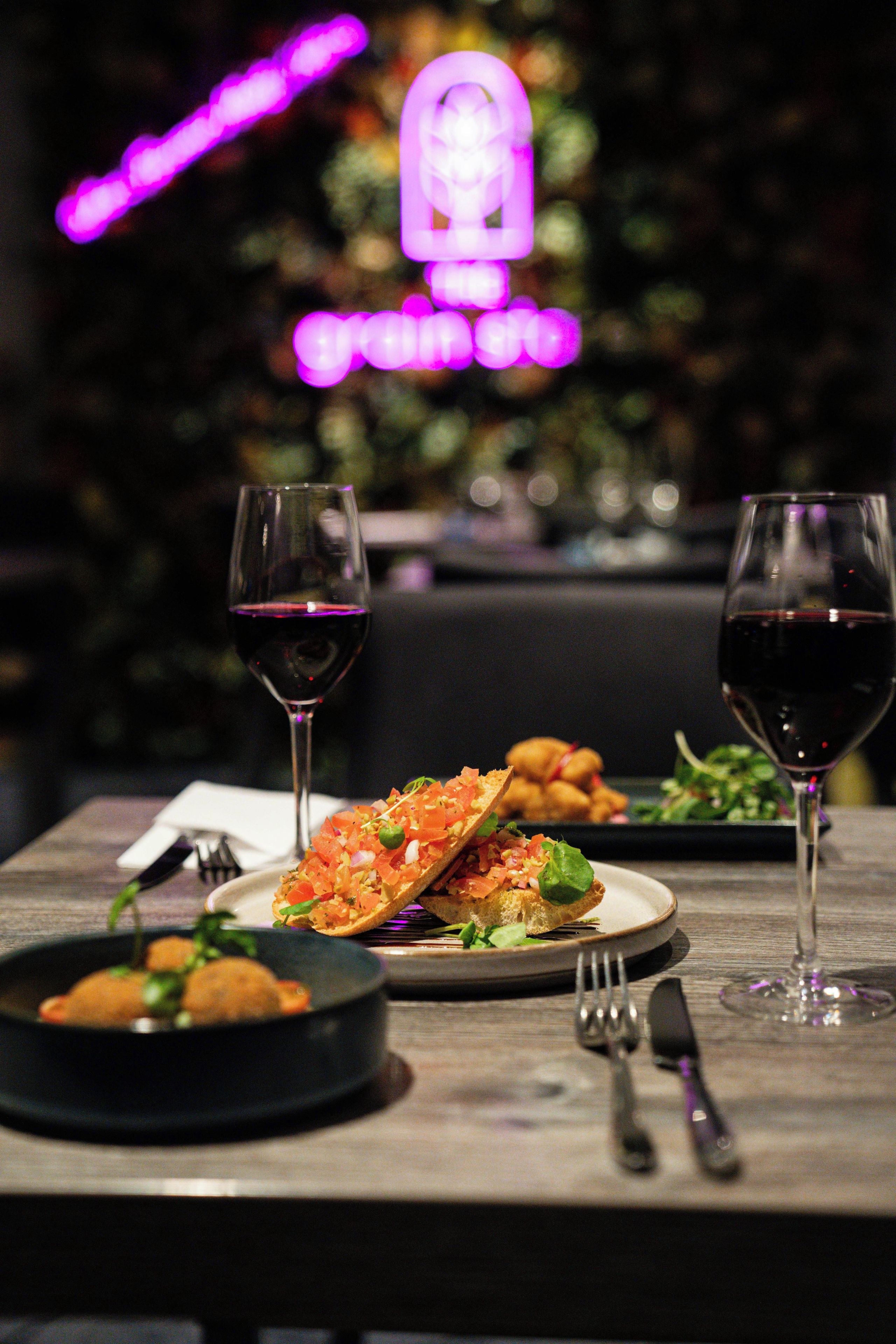 A dining table with two wine glasses, a plate of food, and a neon sign in the background.
