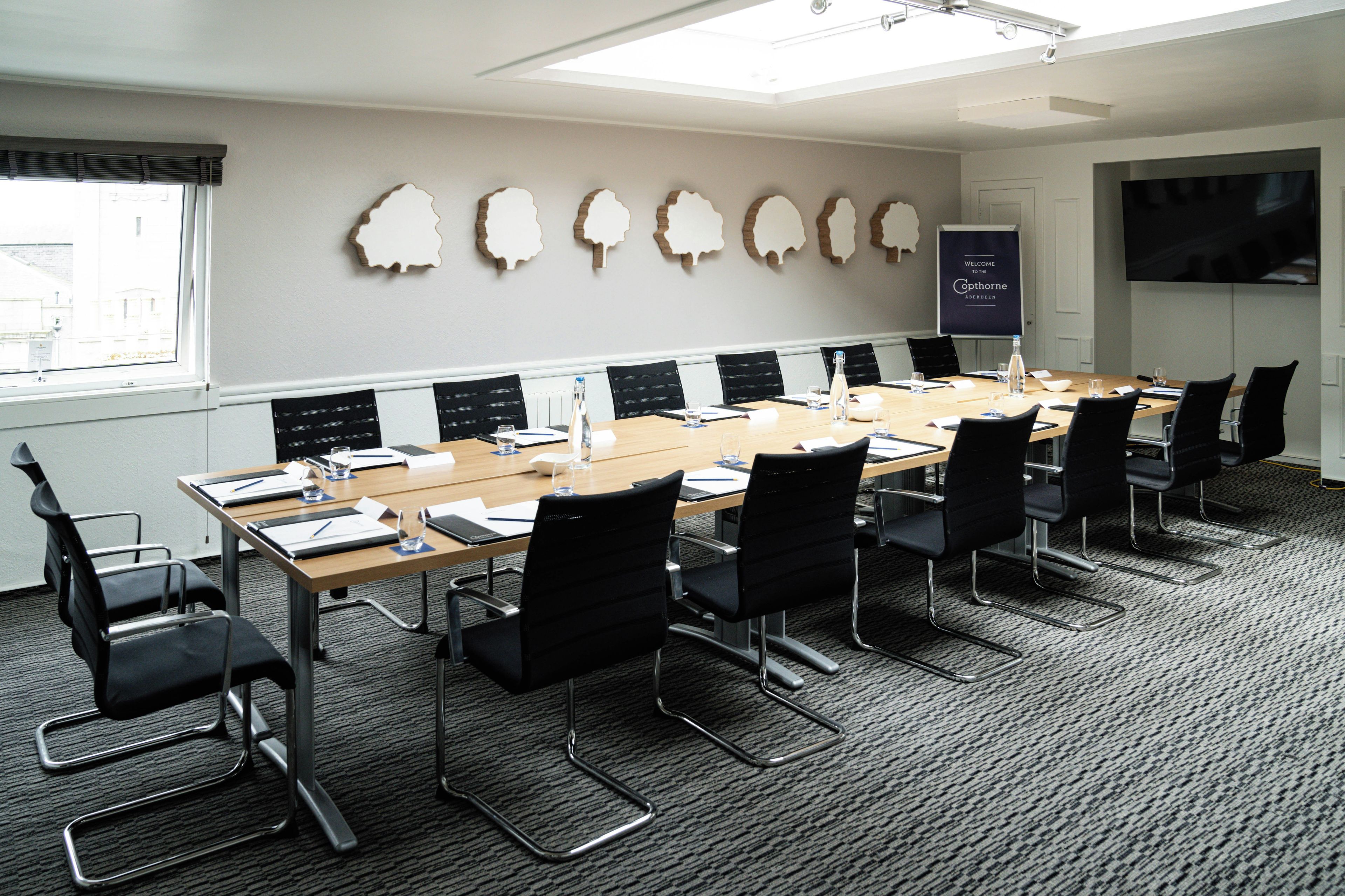 Sala de conferências com uma mesa comprida, cadeiras pretas e tela de projetor.