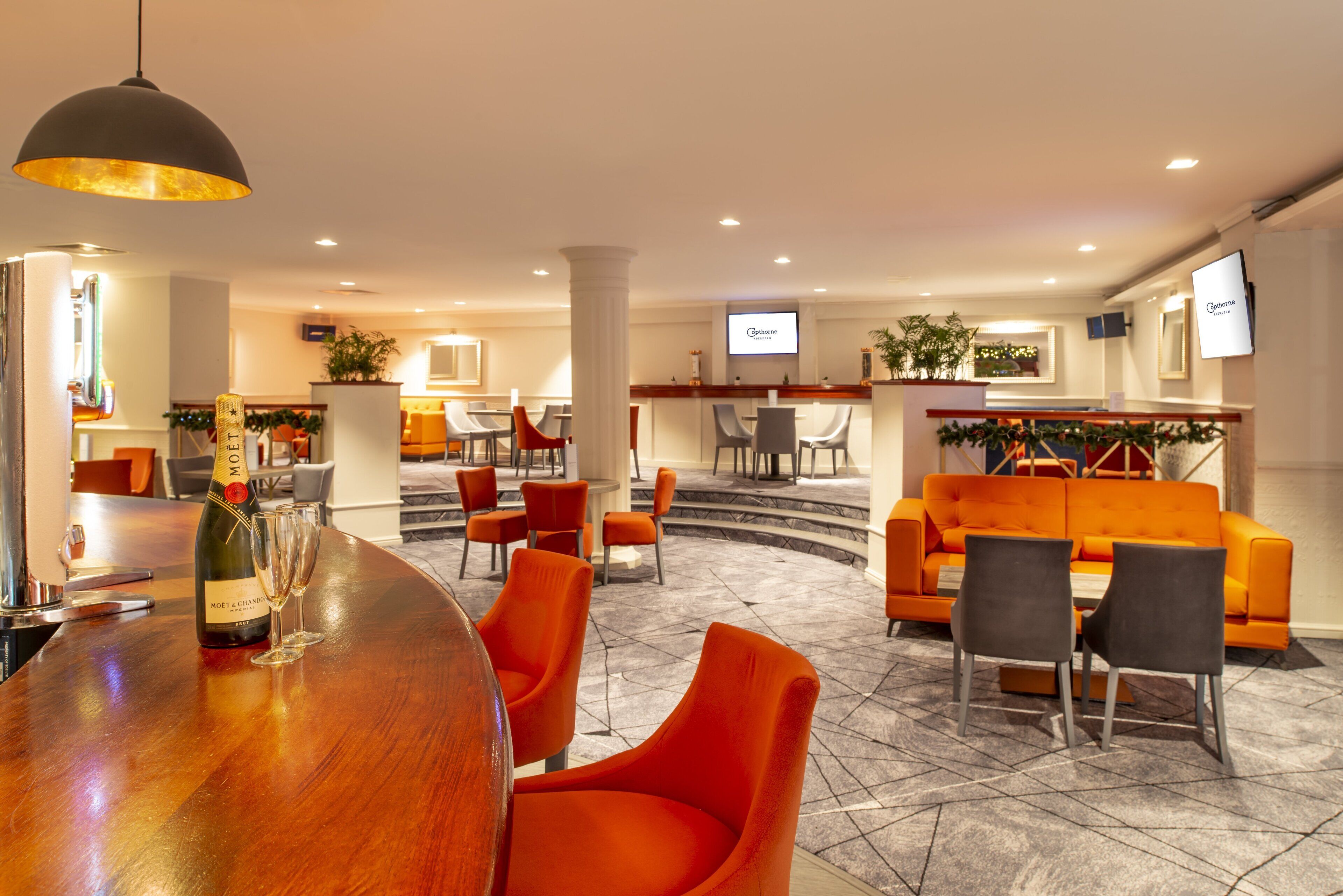 Um hall de hotel com área de bar, assentos laranja, uma escadaria e uma tela de televisão exibindo o nome do hotel.