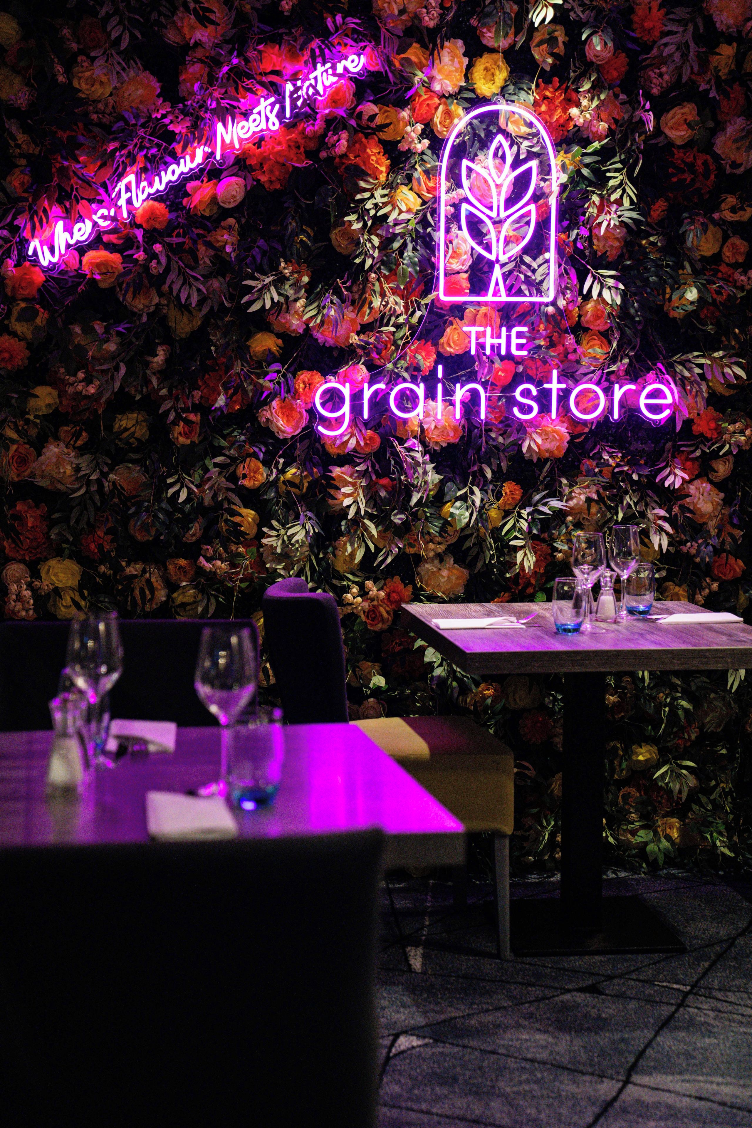 A restaurant interior with a neon sign reading 'The Grain Store' and a floral wall backdrop.