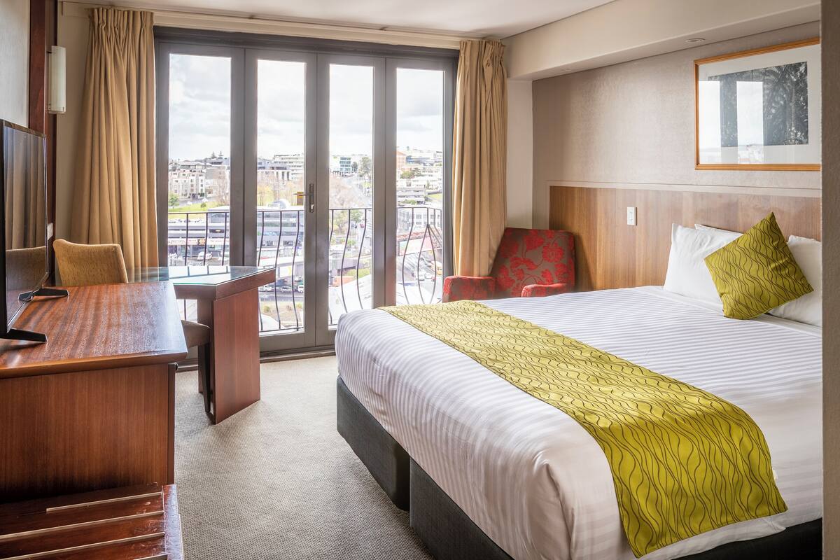 A hotel room with a large bed, a desk, a chair, and a view of the city through large windows.