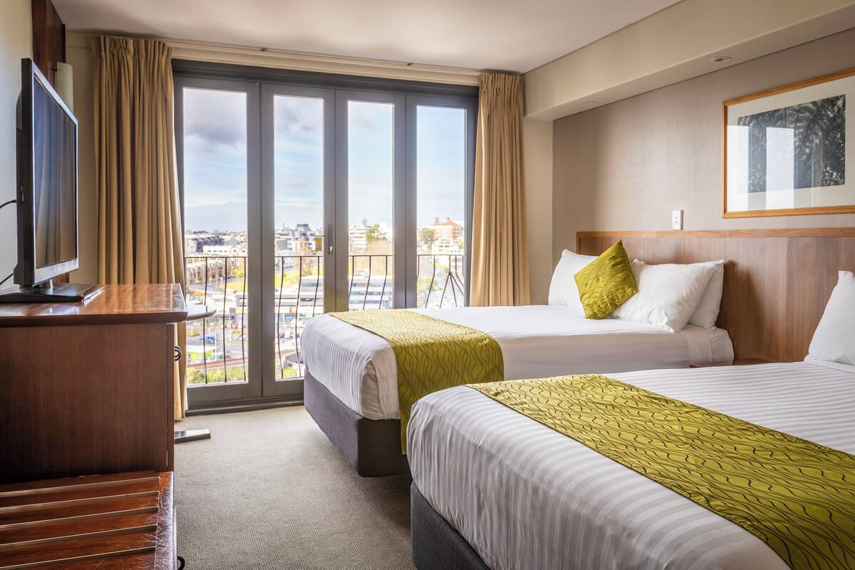 A hotel room with two beds, a TV, and a balcony with a city view.
