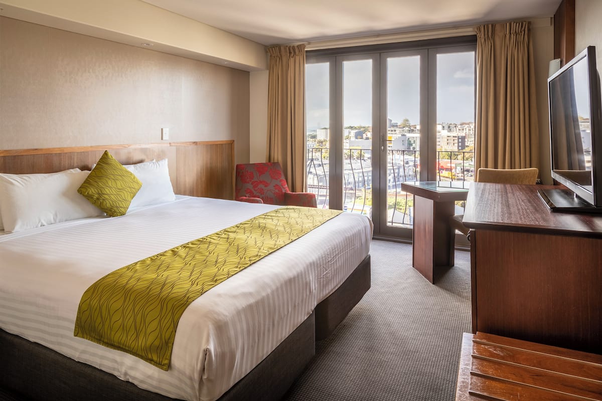 A hotel room with a large bed, a desk, a TV, and a balcony with a city view.