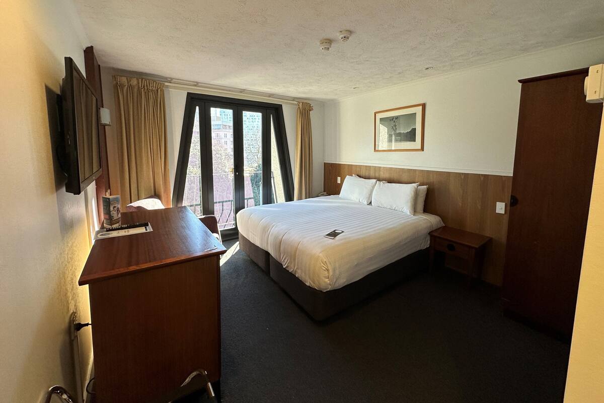 A hotel room with a bed, a desk, a chair, a TV, and a window with curtains.