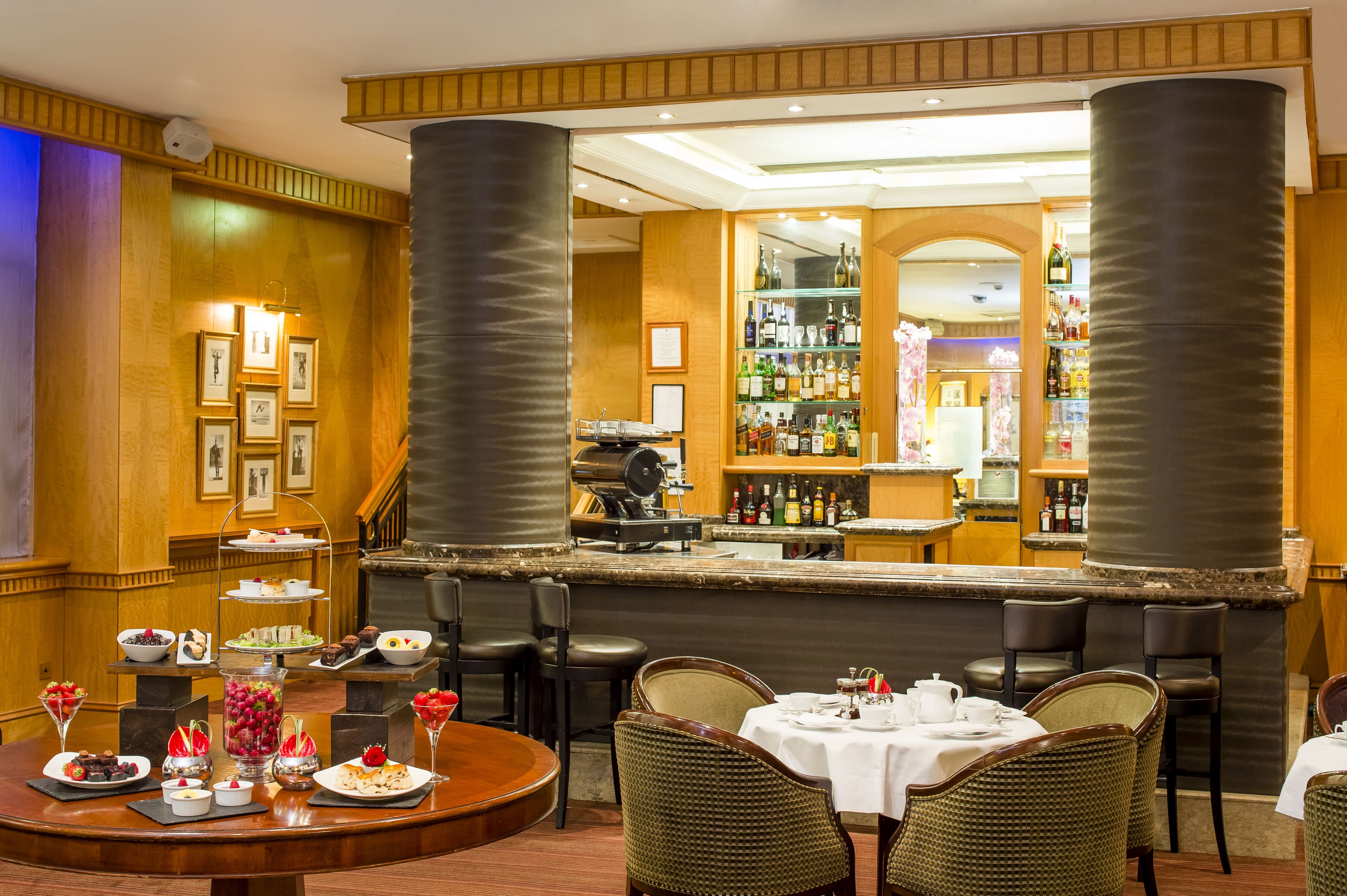 A bar area with a counter, bar stools, and a display of bottles. There are framed pictures on the wall and a buffet table with various dishes.