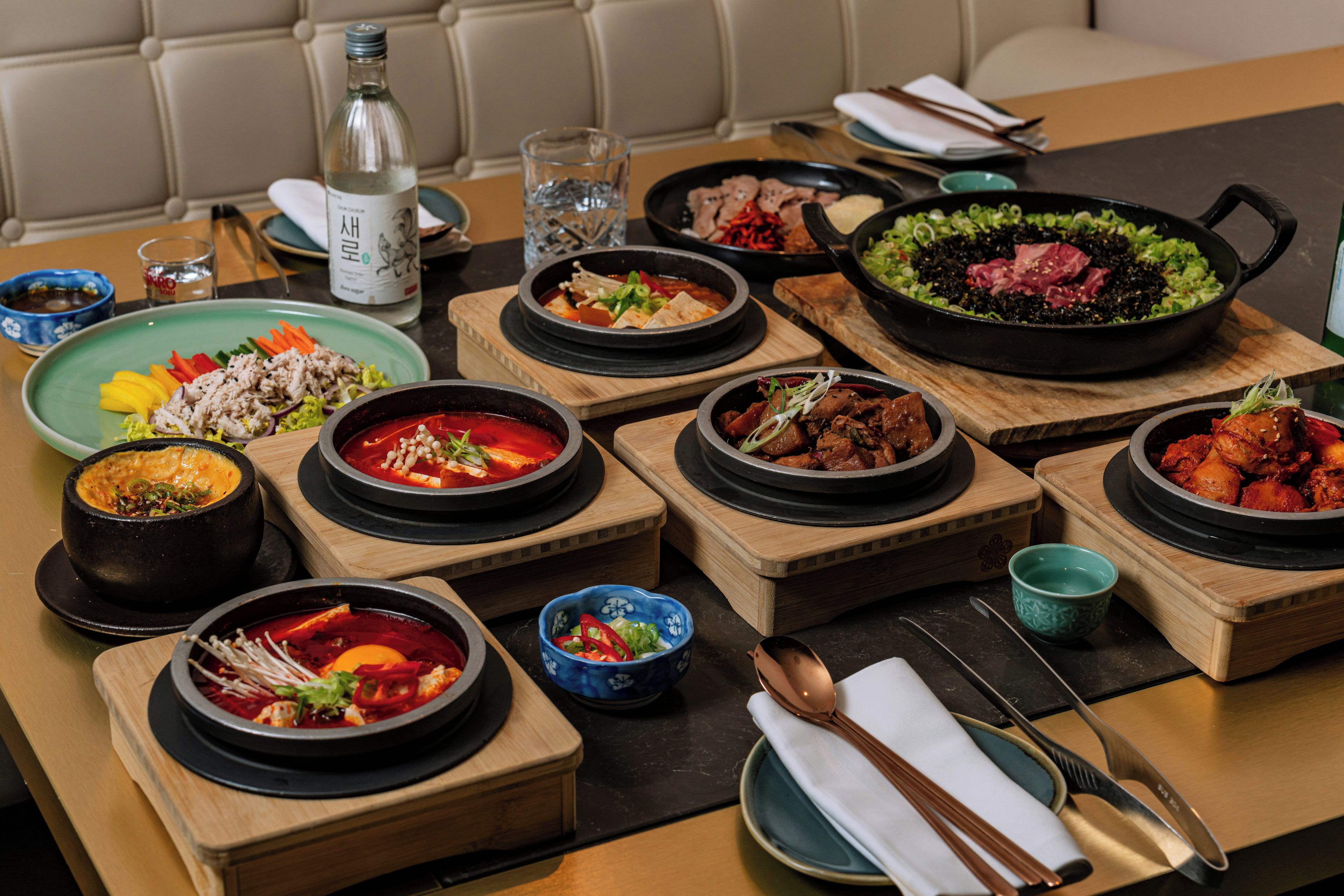 A table set with various Korean dishes, including pots of soup and grilled meat, accompanied by bowls of side dishes and a bottle of water.