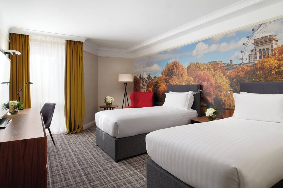 Habitación de hotel con dos camas, un escritorio, una silla, una lámpara y un mural con una vista urbana.