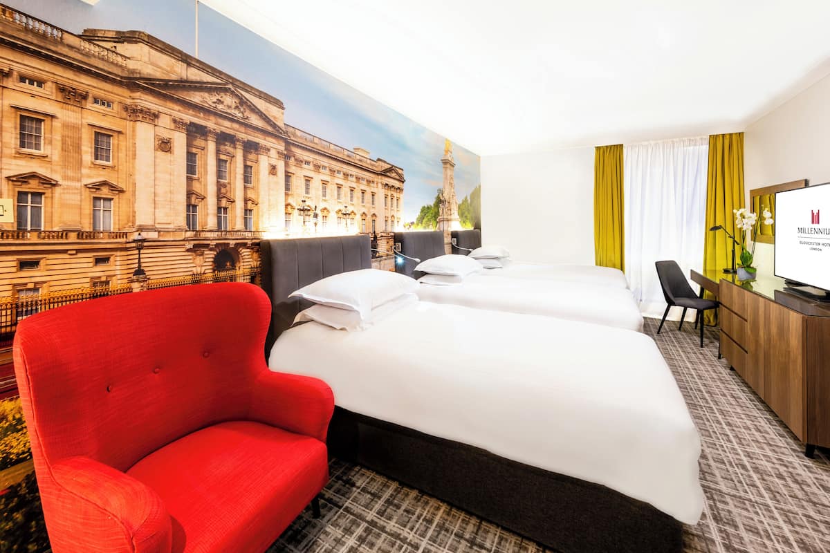 Habitación de hotel con dos camas, un sillón rojo, un escritorio con una computadora y un gran mural de un edificio histórico.