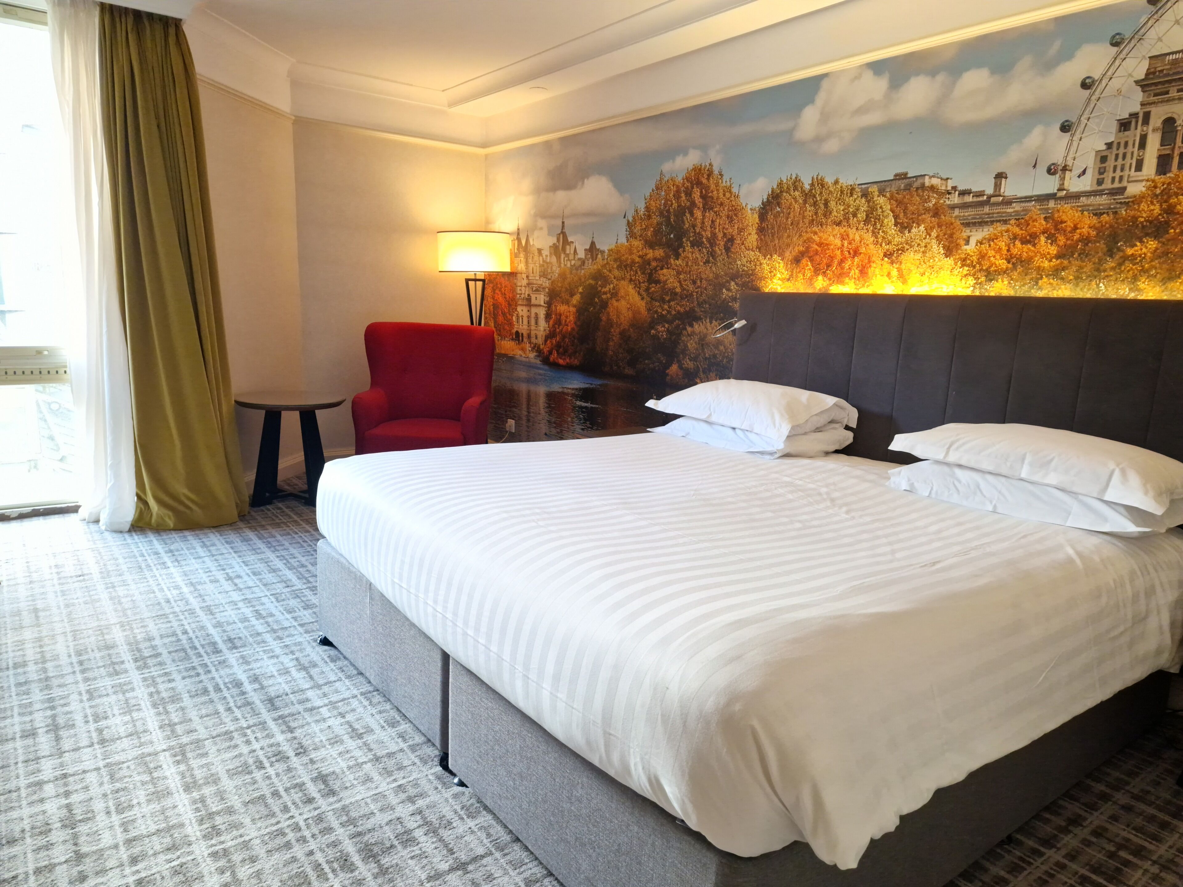 Una habitación de hotel con una cama grande, un sillón rojo, una mesita pequeña y un mural con un paisaje urbano en la pared.