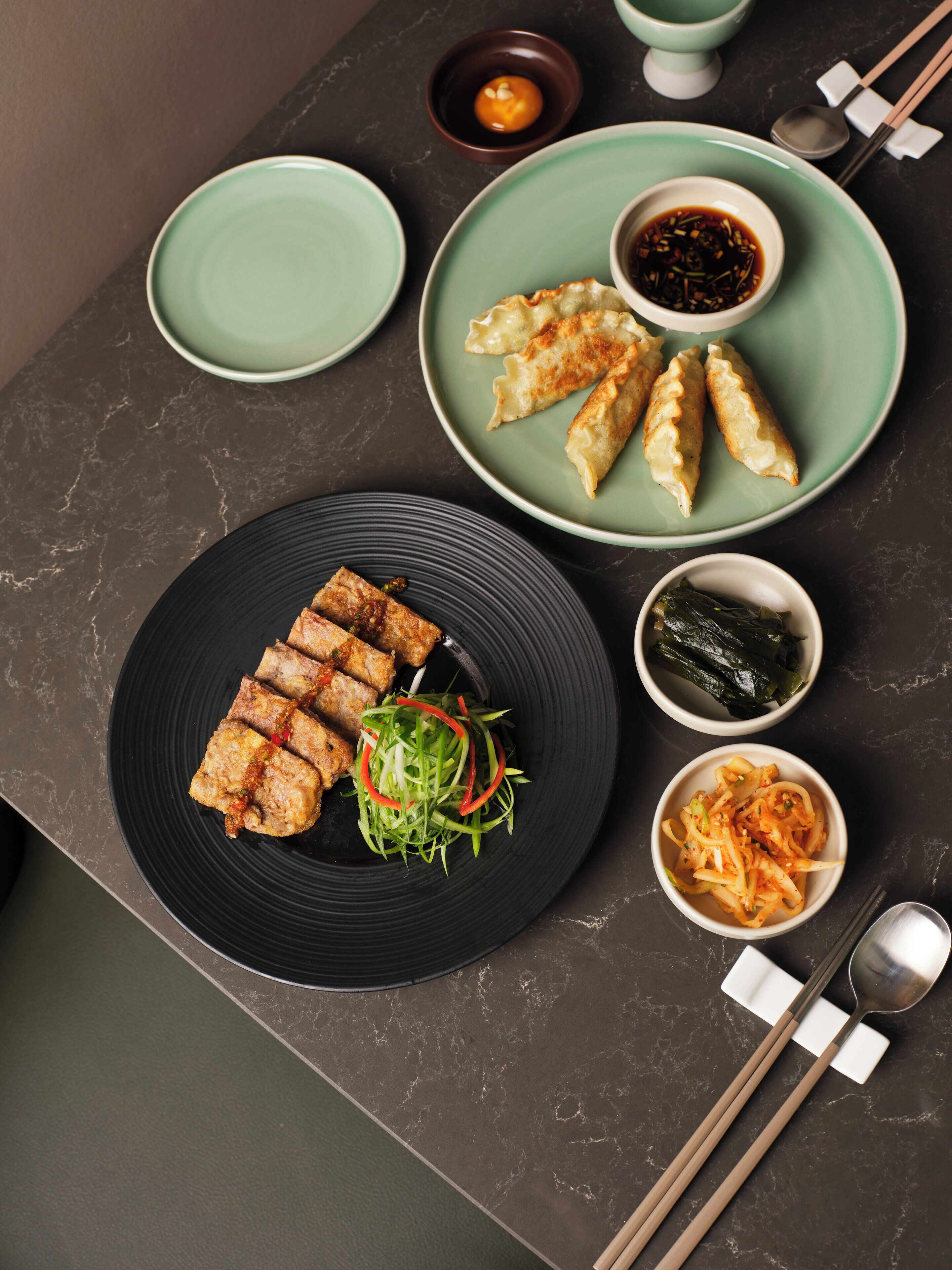 A table with various dishes, including dumplings and a salad, accompanied by dipping sauces and seaweed.