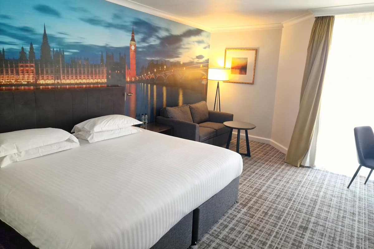 Habitación de hotel con una cama grande, un sofá, una mesita, una lámpara y un mural con vista a la ciudad.