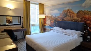 Quarto de hotel com uma cama grande, uma mesa de trabalho com cadeira, um espelho e uma janela com cortinas.