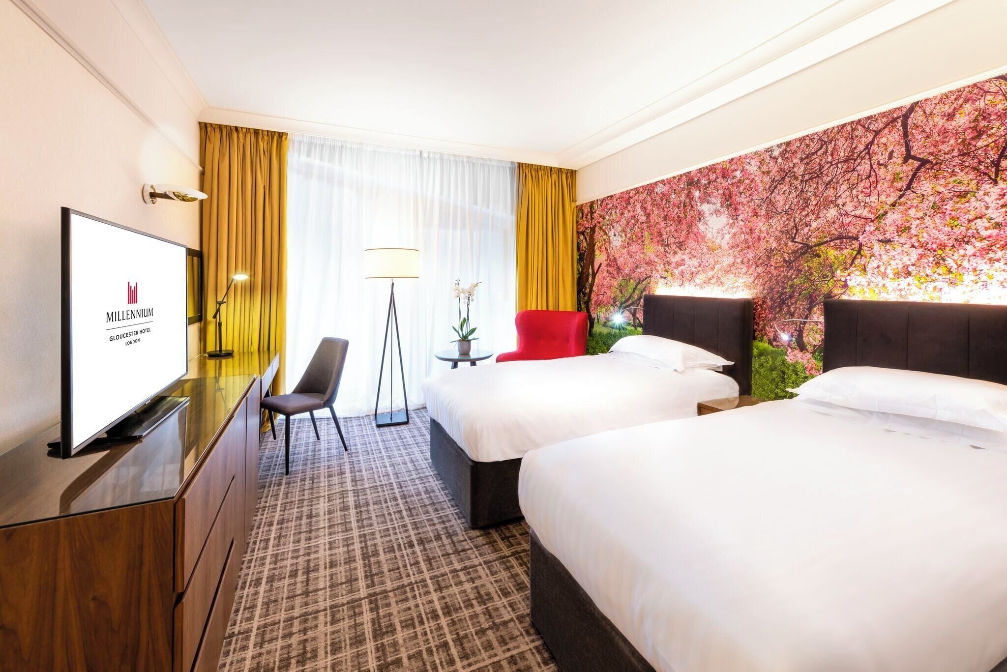A hotel room with two beds, a desk with a TV, a chair, a lamp, and a large floral wall mural.