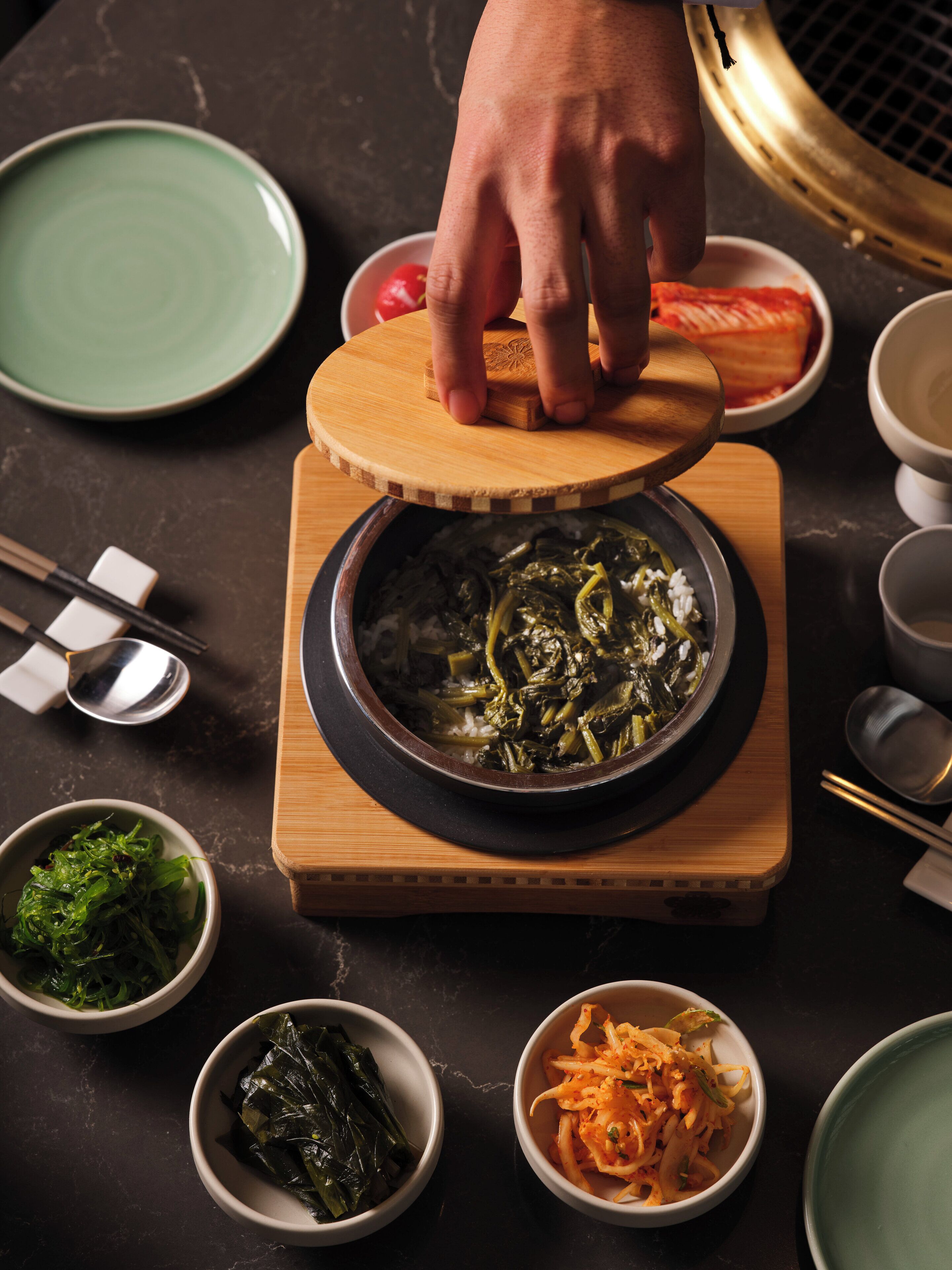 Una mesa con un recipiente de cocción caliente, palillos, tazones de verduras y una mano levantando una tapa.