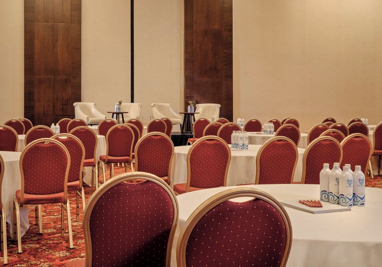 Un salón de conferencias con filas de sillas tapizadas en rojo y mesas blancas, equipado con botellas de agua.