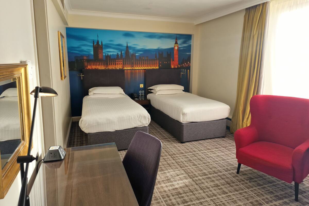 Habitación de hotel con dos camas individuales, un escritorio con lámpara, un sillón rojo y un gran mural en la pared con una vista urbana.