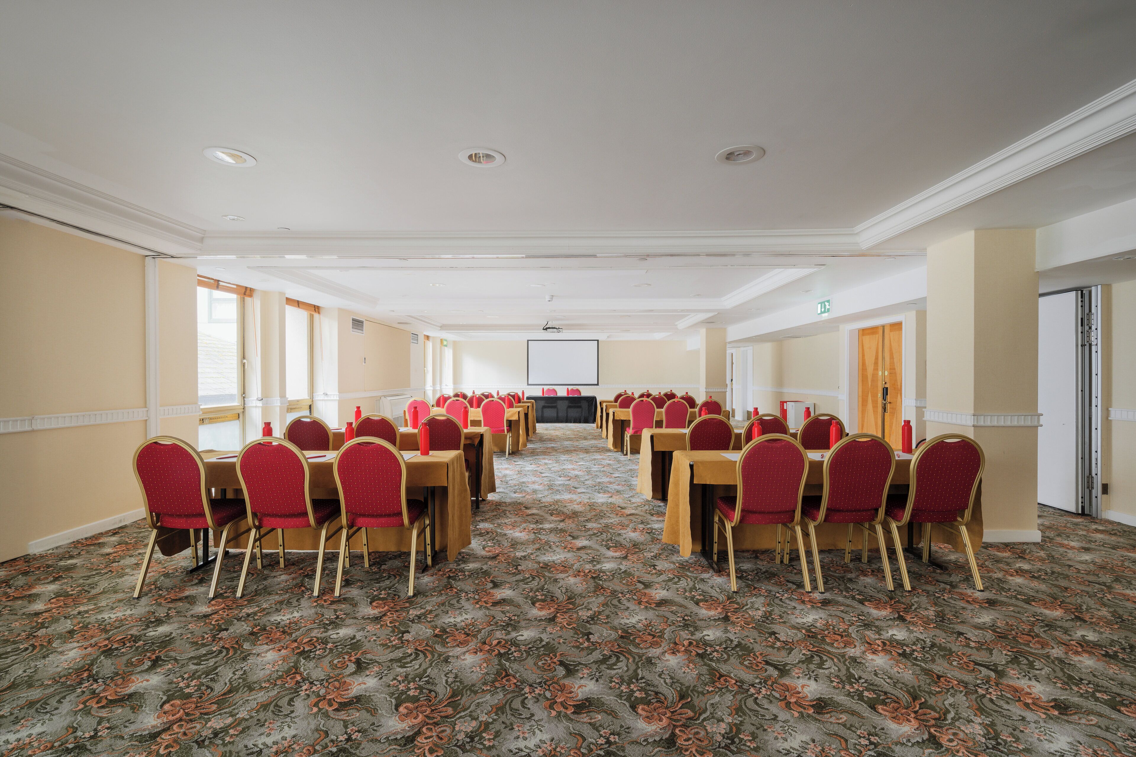A conference room with rows of tables and chairs, a carpeted floor, and a projector screen at the front.