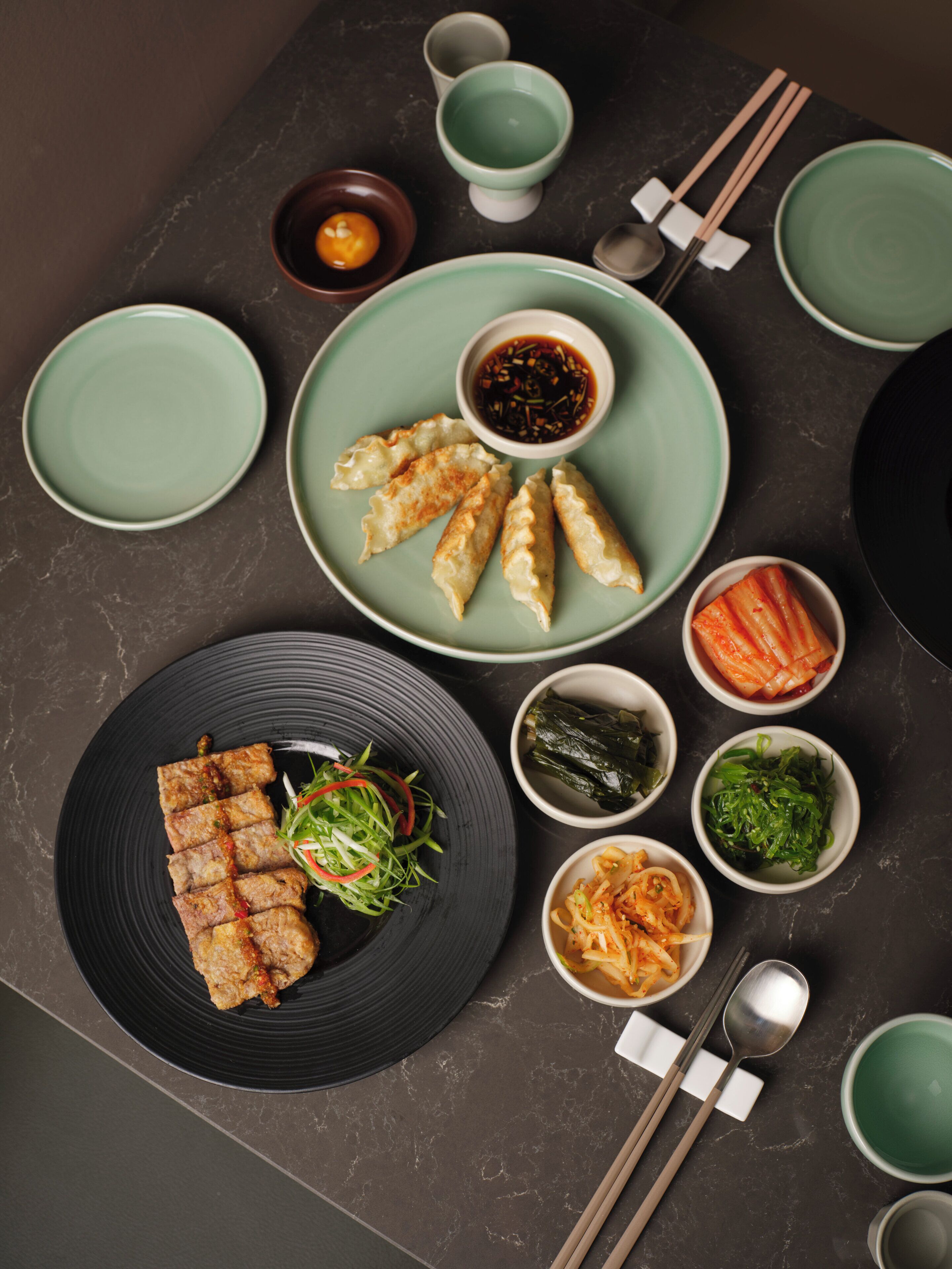 Una mesa con diversos platos, como dumplings, kimchi y una ensalada, junto con palillos y tazones.