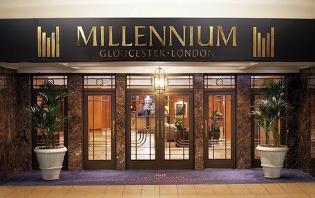 The exterior of Millennium Gloucester London, featuring a grand entrance with a large sign, two potted plants, and a well-lit interior visible through the glass doors.
