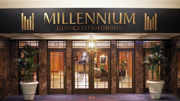 Das Äußere des Millennium Gloucester London mit einem prunkvollen Eingang, einem großen Schild, zwei Topfpflanzen und einem gut beleuchteten Innenraum, der durch die Glasüren sichtbar ist.