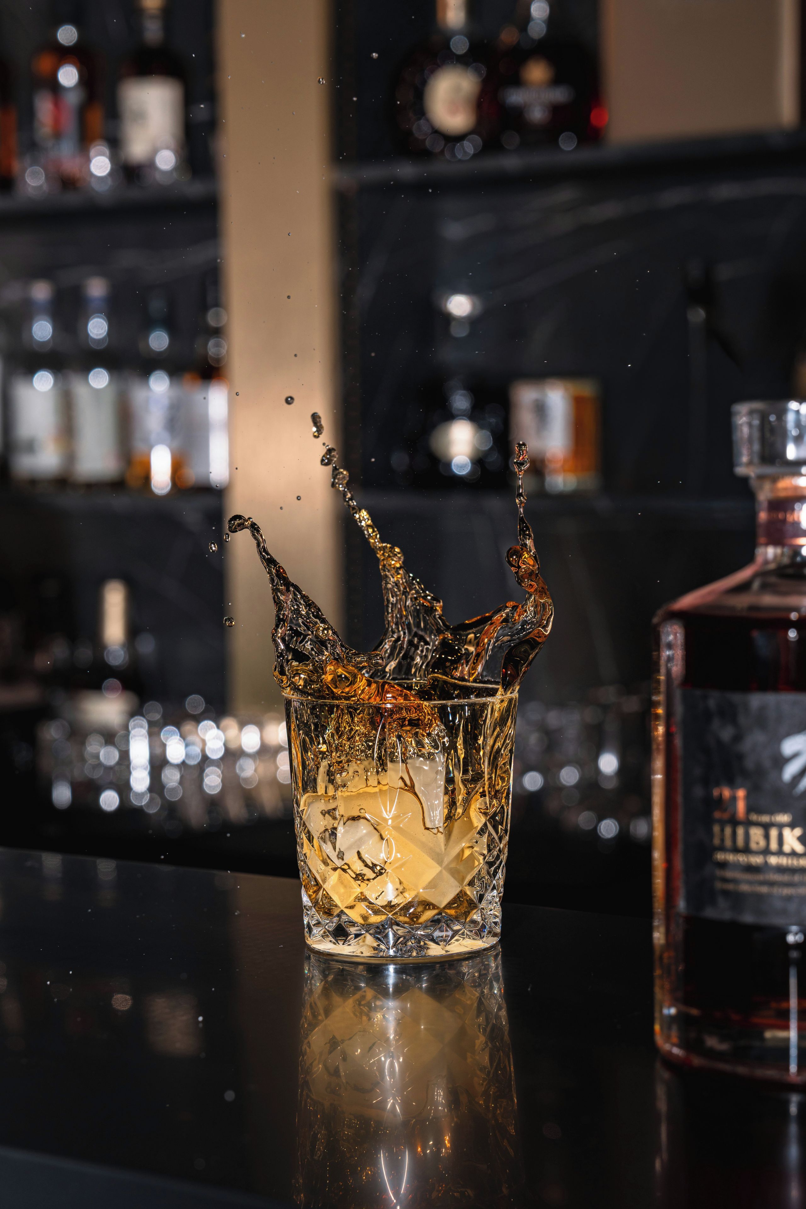 Un vaso con un poco de líquido sobre una superficie reflectante, una botella de licor y un fondo borroso con estantes.