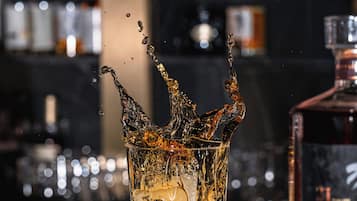 Un verre contenant un peu de liquide, posé sur une surface réfléchissante, une bouteille de spiritueux et un arrière-plan flou avec des étagères.