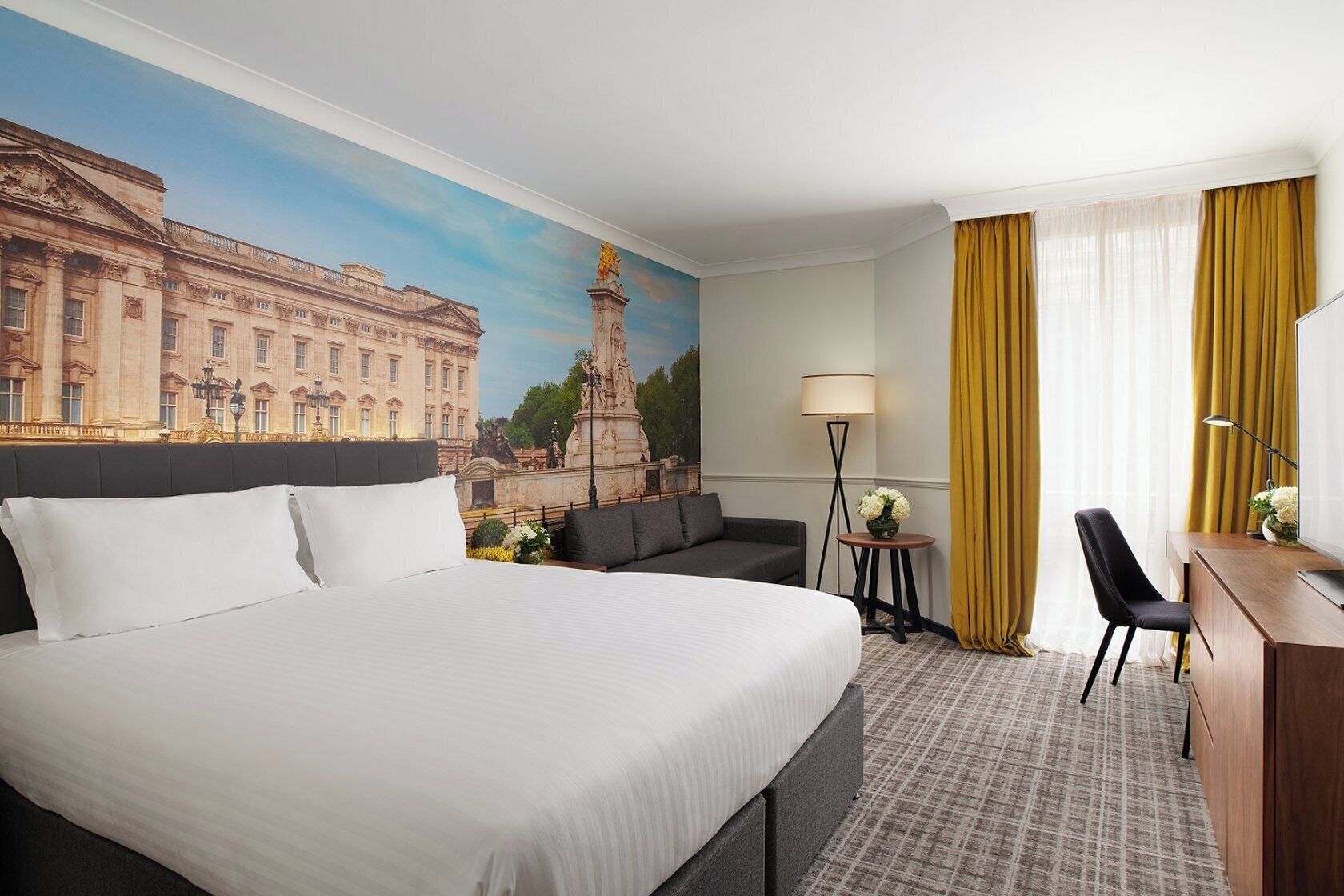 Une chambre d’hôtel avec un grand lit, un canapé, un bureau et une lampe. Un tableau représentant un bâtiment est accroché au mur.