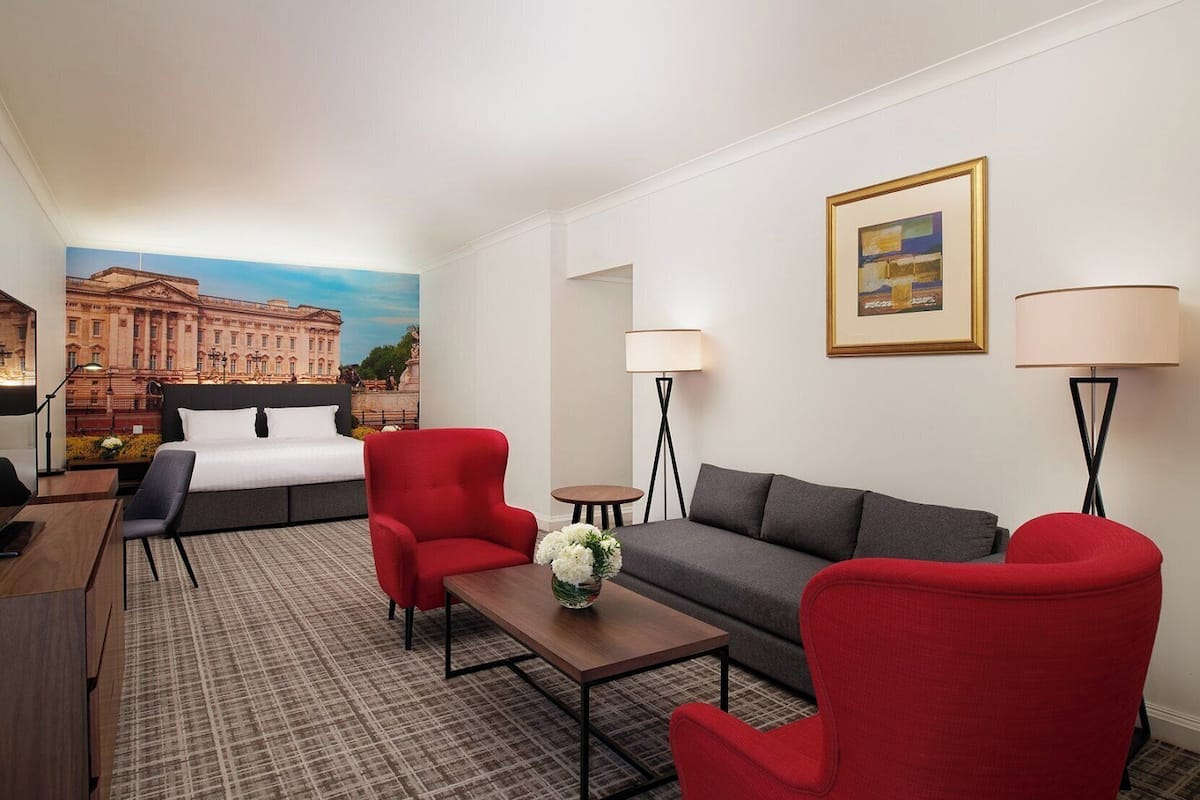 Una habitación de hotel con una cama, dos sillones rojos, un sofá gris, una mesita con un jarrón, dos lámparas de pie y un cuadro en la pared.