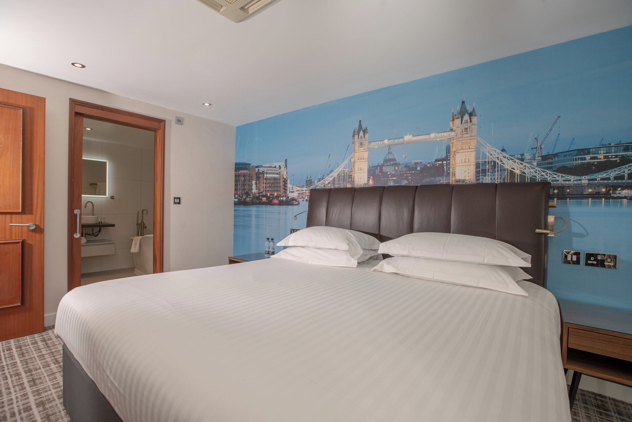 A hotel room with a large bed, a view of the Tower Bridge, and a bathroom visible through an open door.