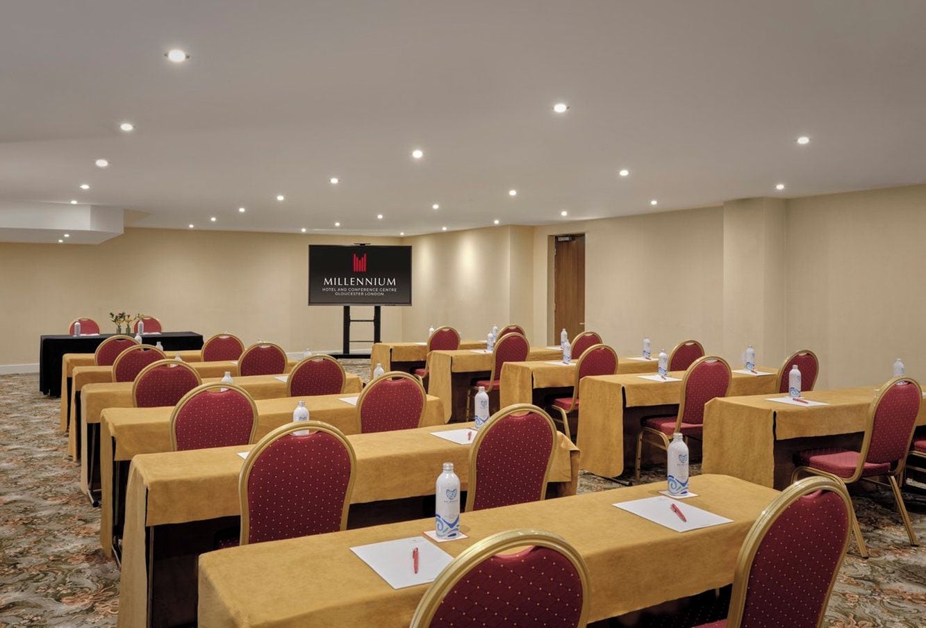 A conference room with rows of tables and chairs, a screen displaying the hotel name, and a carpeted floor.