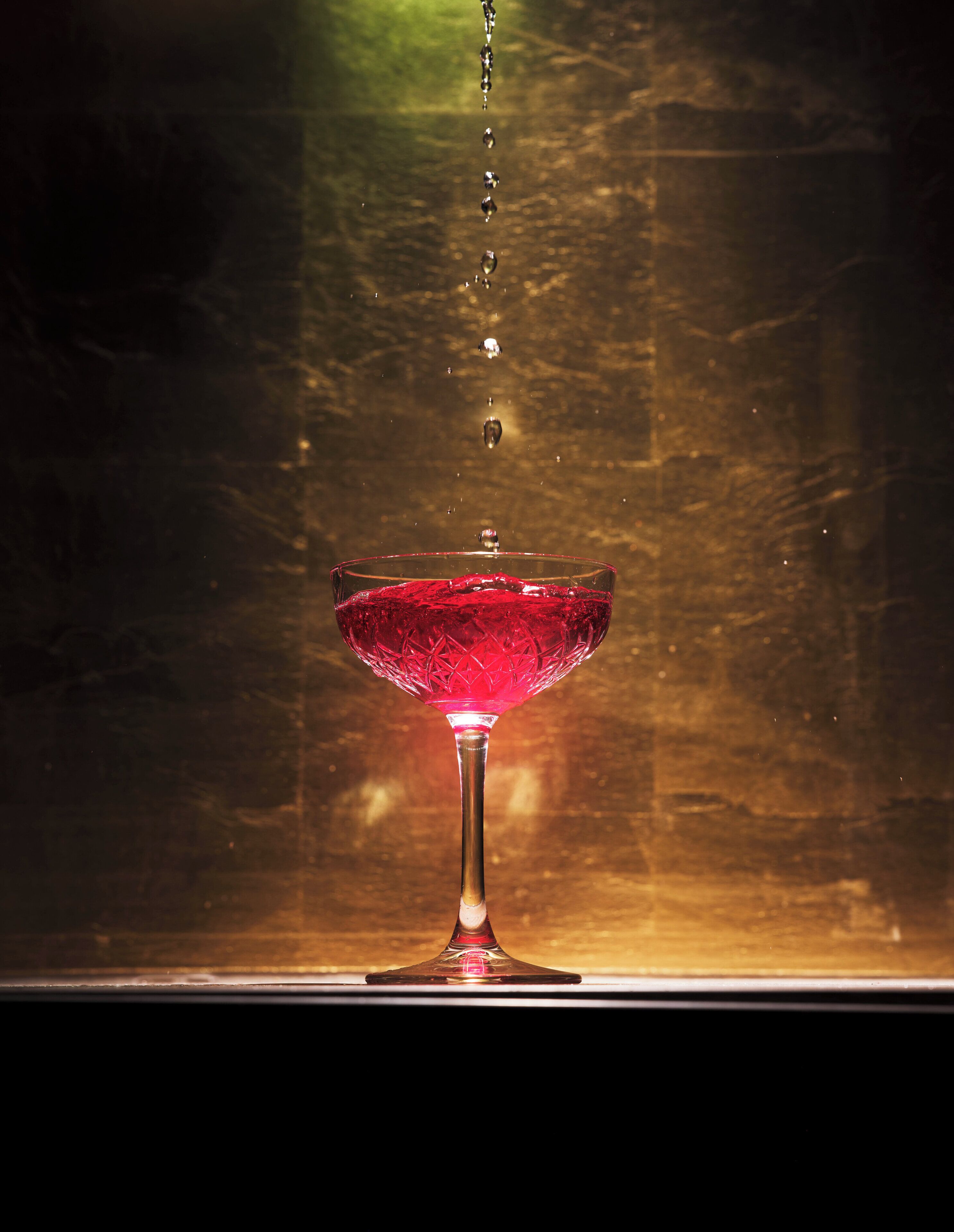 Ein klares Glas mit einer roten Flüssigkeit, möglicherweise ein Cocktail, bei dem Wassertropfen hineinfallen.