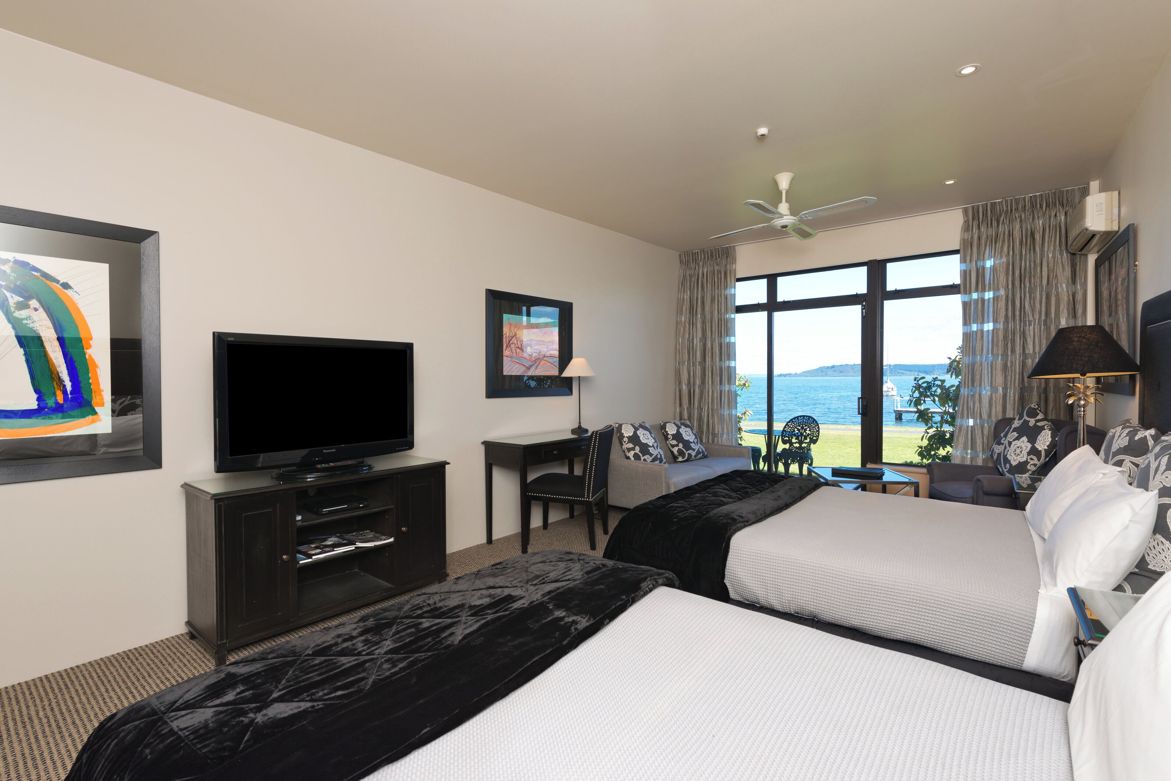 A hotel room with two beds, a TV, a desk, and a view of the sea.