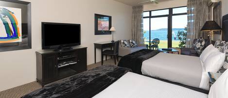A hotel room with two beds, a TV, a desk, and a view of the sea.