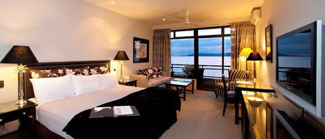 A hotel room with a large bed, a desk, a sofa, a TV, and a view of the sea.