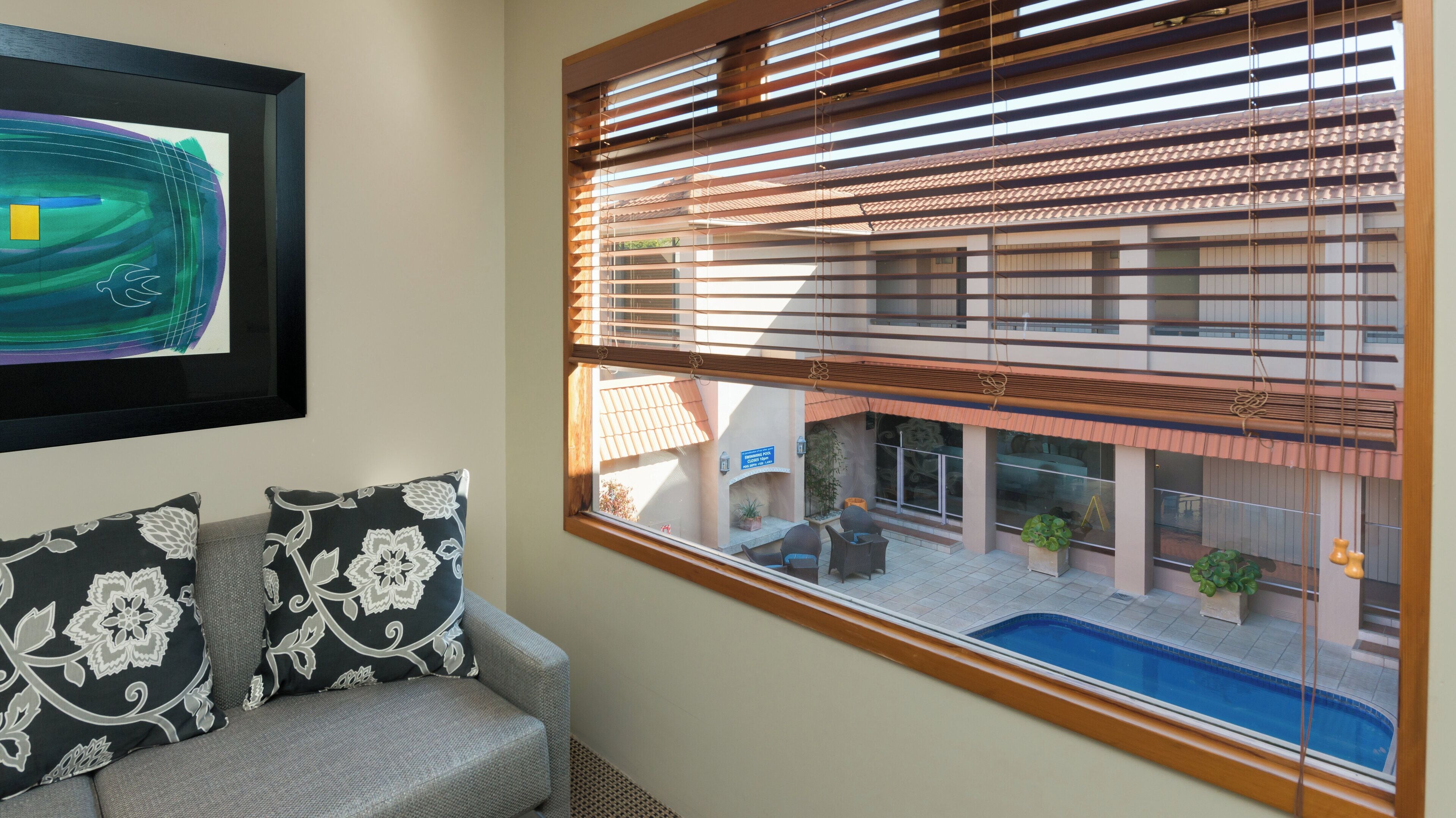 Una habitación con vista a una piscina y un edificio con techo de tejas. Hay un cuadro abstracto enmarcado en la pared y dos almohadones estampados sobre un sofá gris.