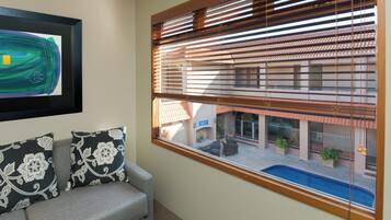 Una habitación con vista a una piscina y un edificio con techo de tejas. Hay un cuadro abstracto enmarcado en la pared y dos almohadones estampados sobre un sofá gris.