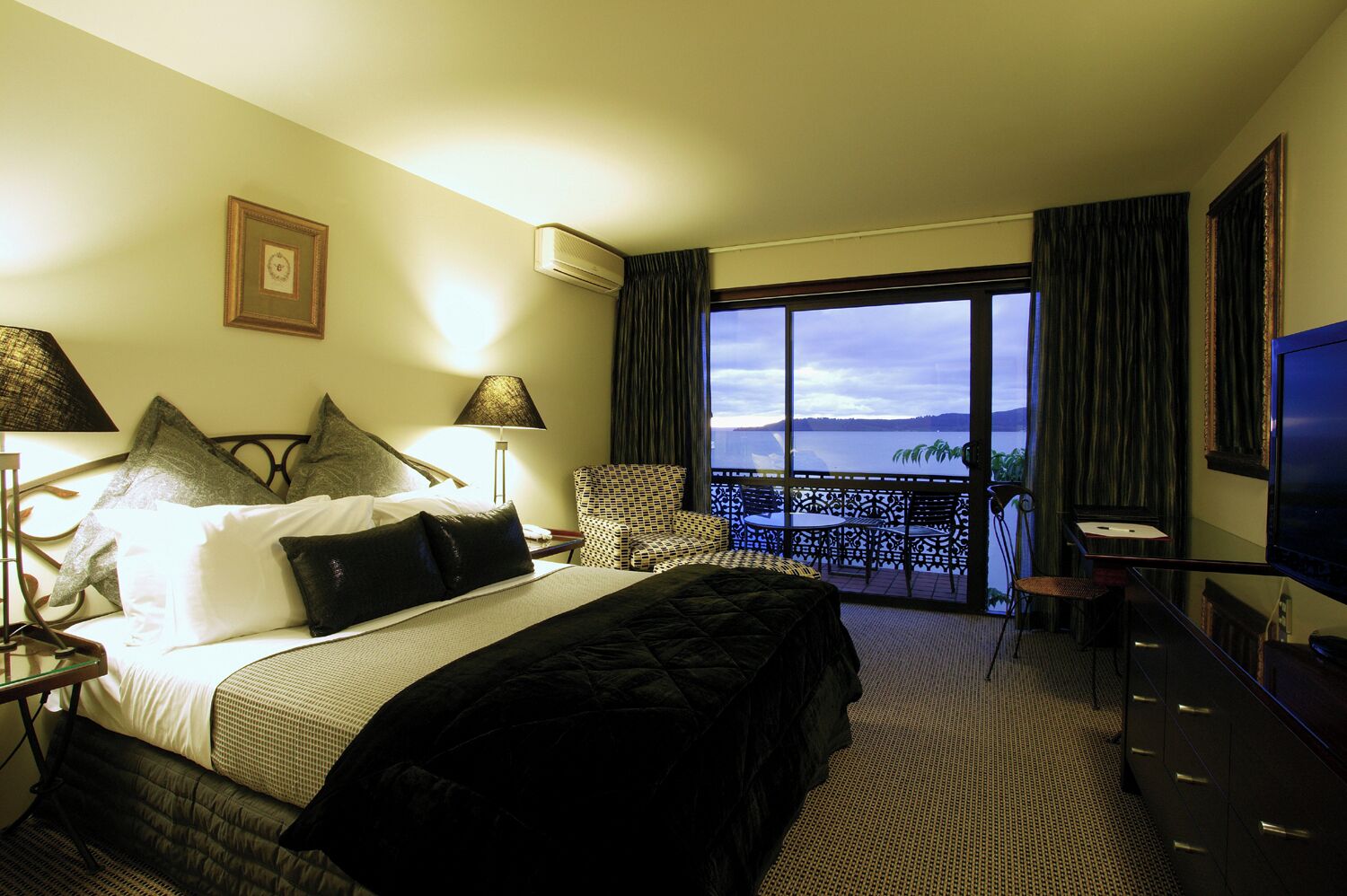 Een hotelkamer met een groot bed, een televisie, een balkon met uitzicht op het water en een bureau met een lamp.