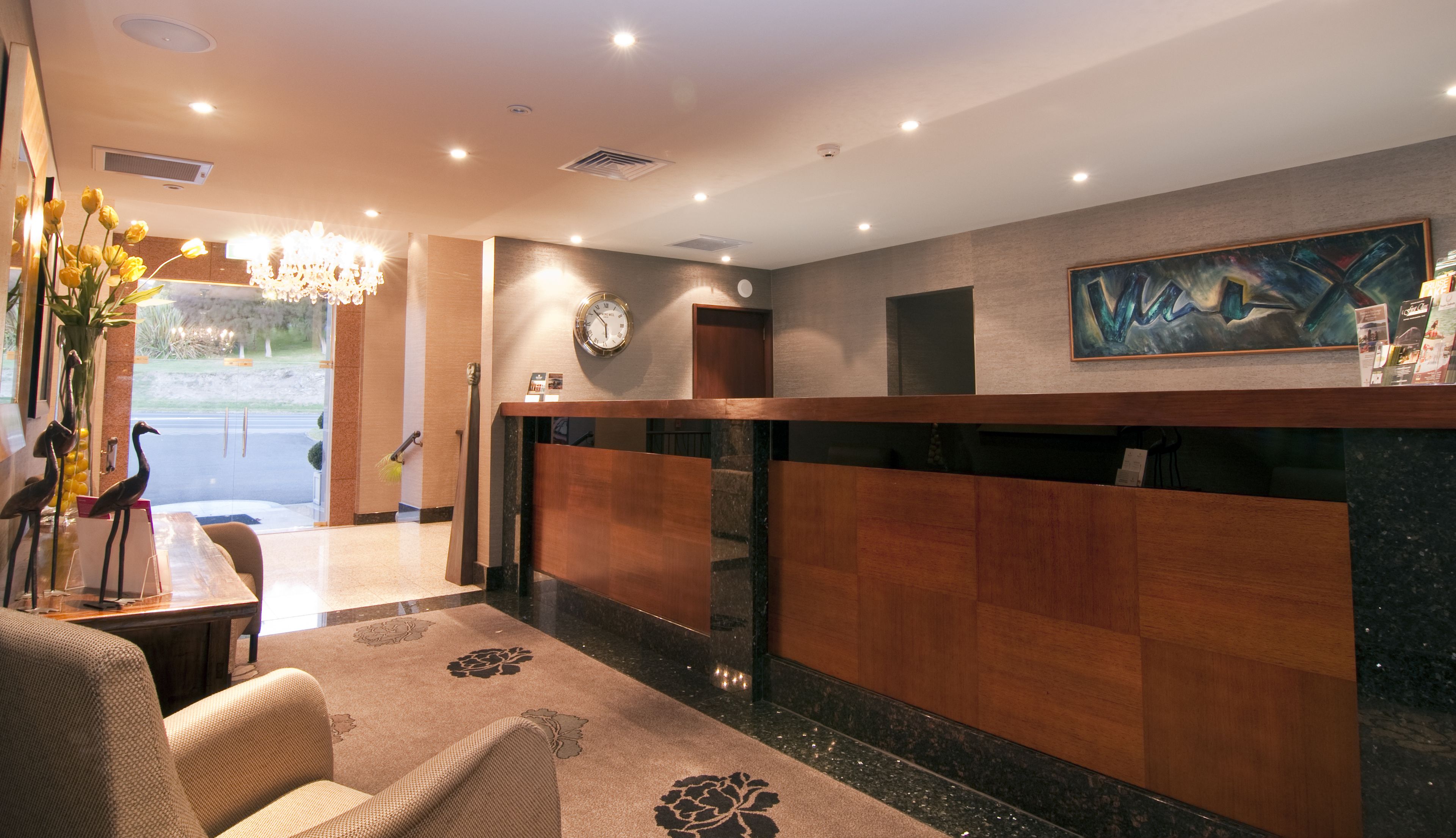 A hotel reception area with a wooden reception desk, a clock, and a painting on the wall.
