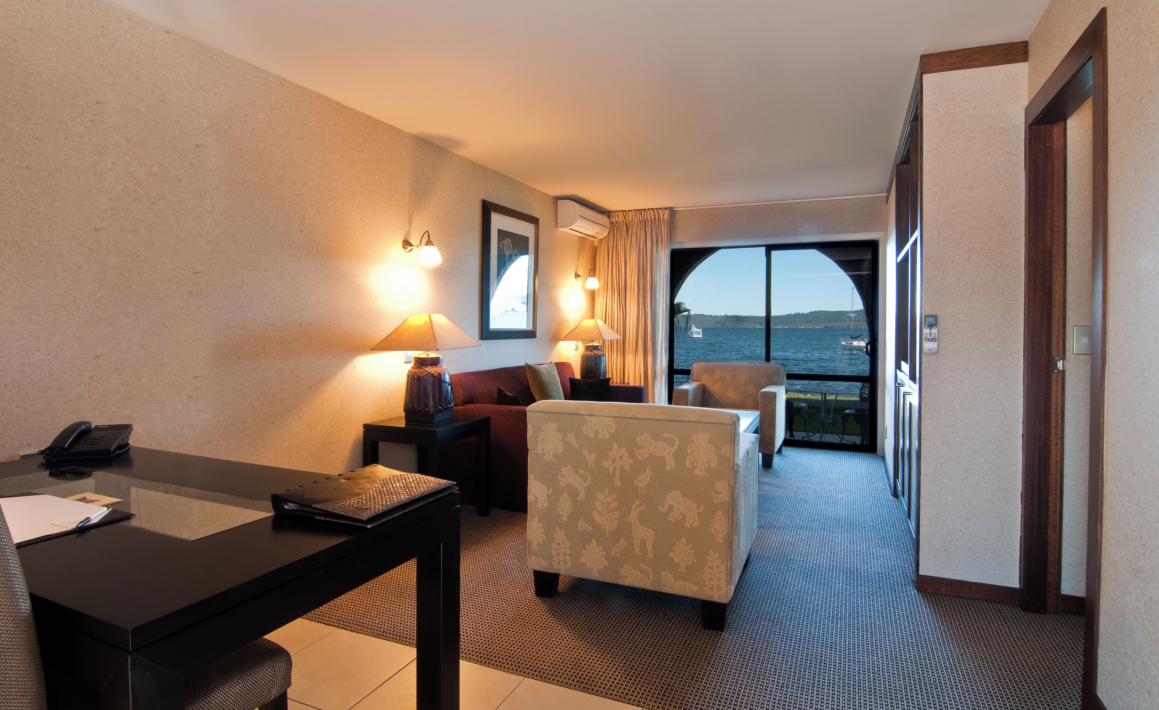 Habitación de hotel con escritorio, sofá, silla y vista al mar a través de un amplio ventanal.
