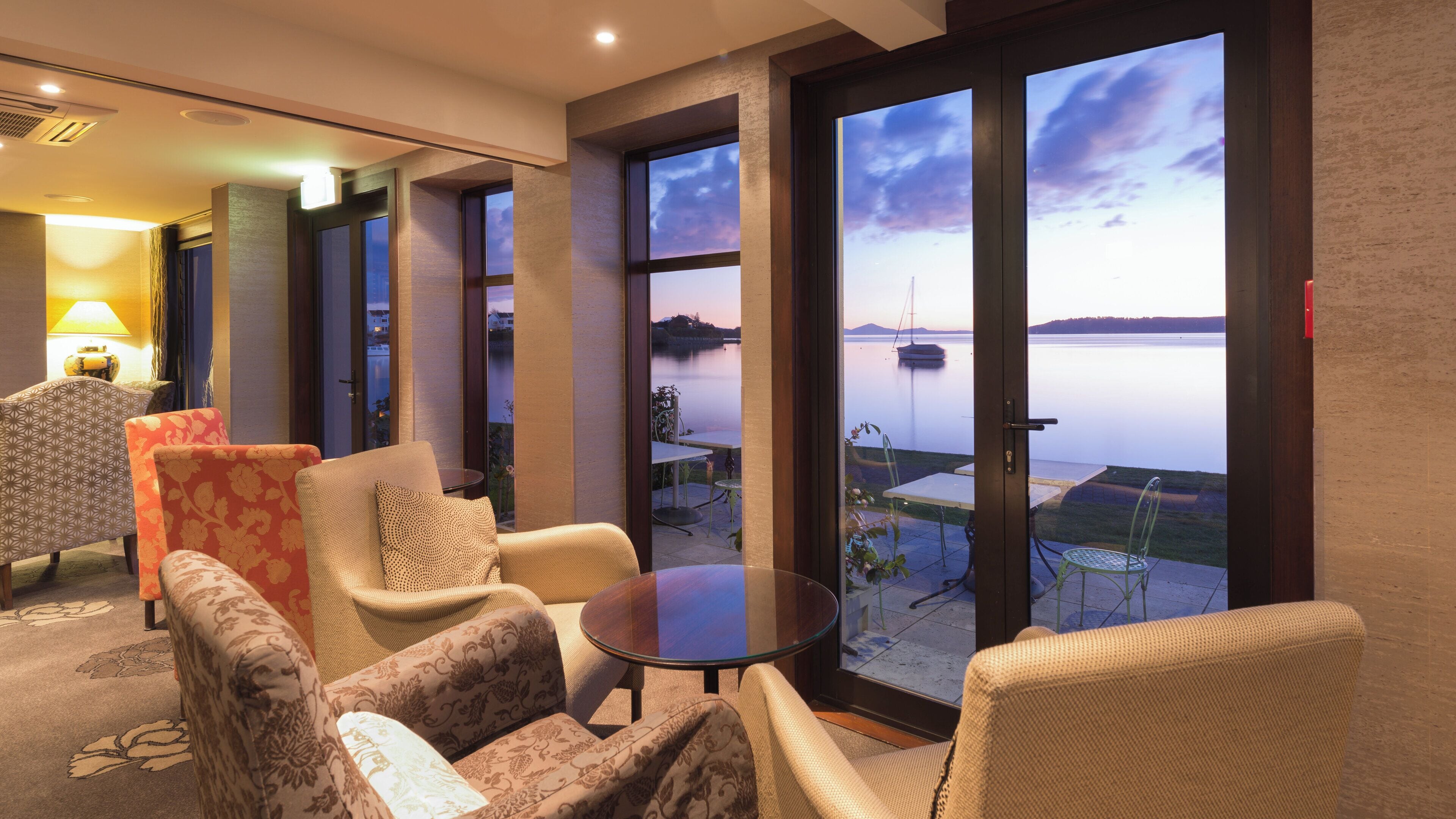 A room with a view of a lake, featuring a table and chairs, a lamp, and a patterned rug.
