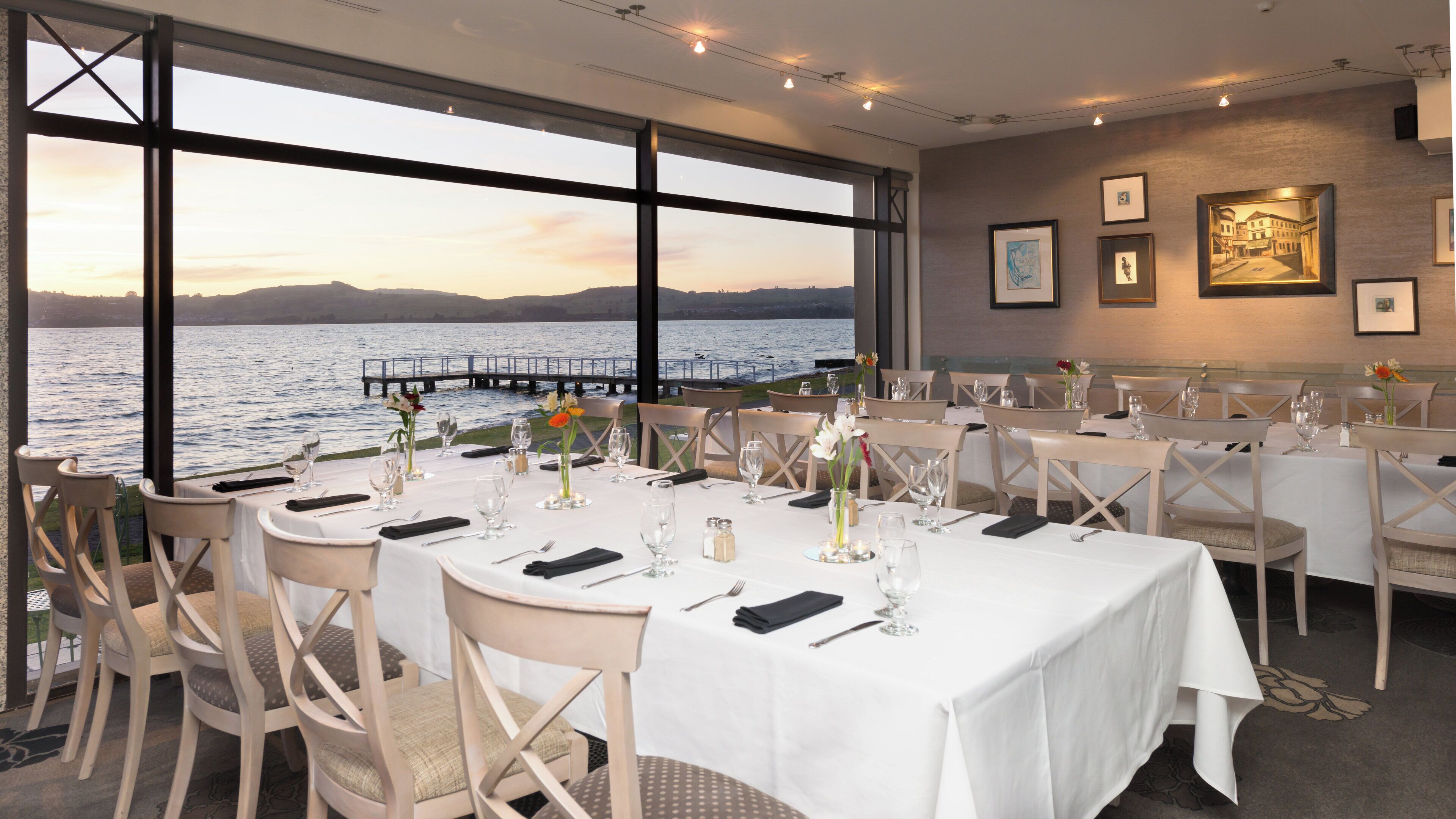 A restaurant with tables set for dining, large windows overlooking a body of water, and framed artwork on the walls.