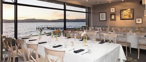 A restaurant with tables set for dining, large windows overlooking a body of water, and framed artwork on the walls.