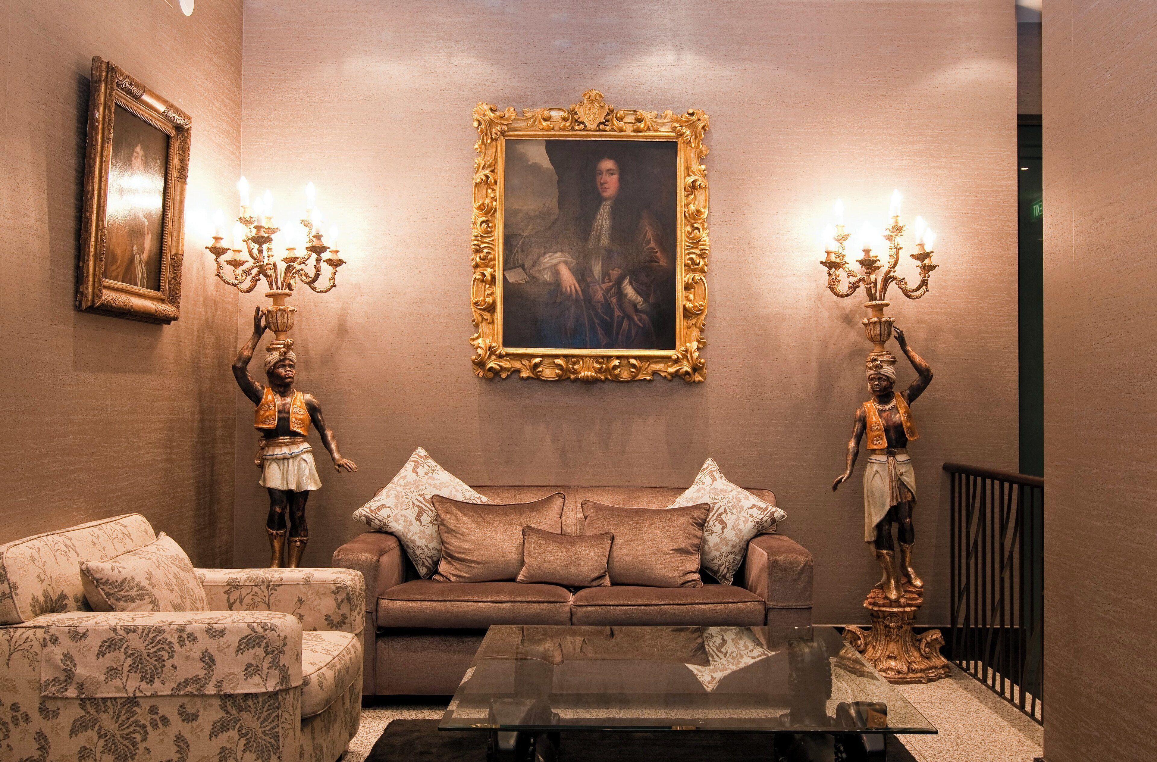 Una stanza con un divano marrone, due poltrone, un tavolino in vetro, un ritratto incorniciato e due elaborati candelabri.
