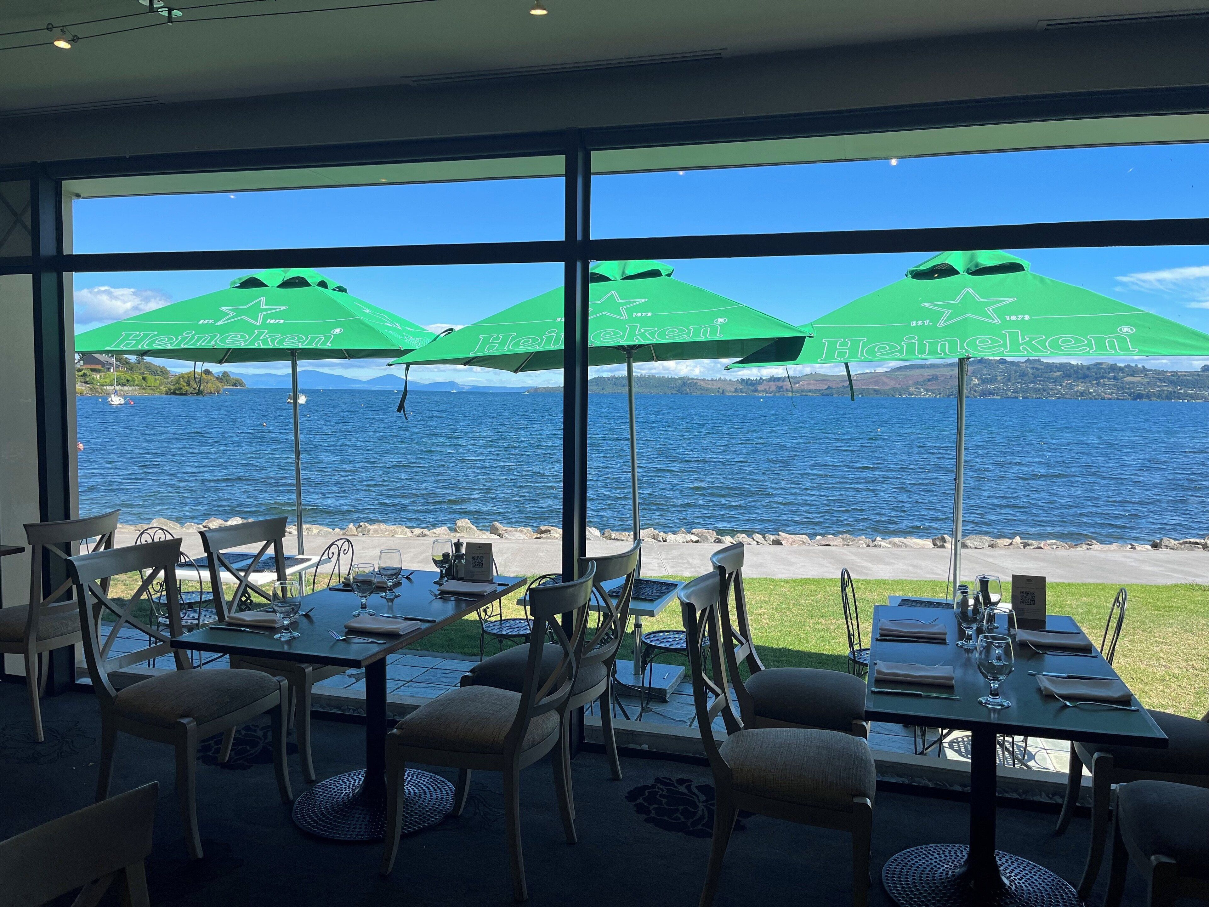 A restaurant with large windows overlooking a lake, featuring green umbrellas with Heineken branding.