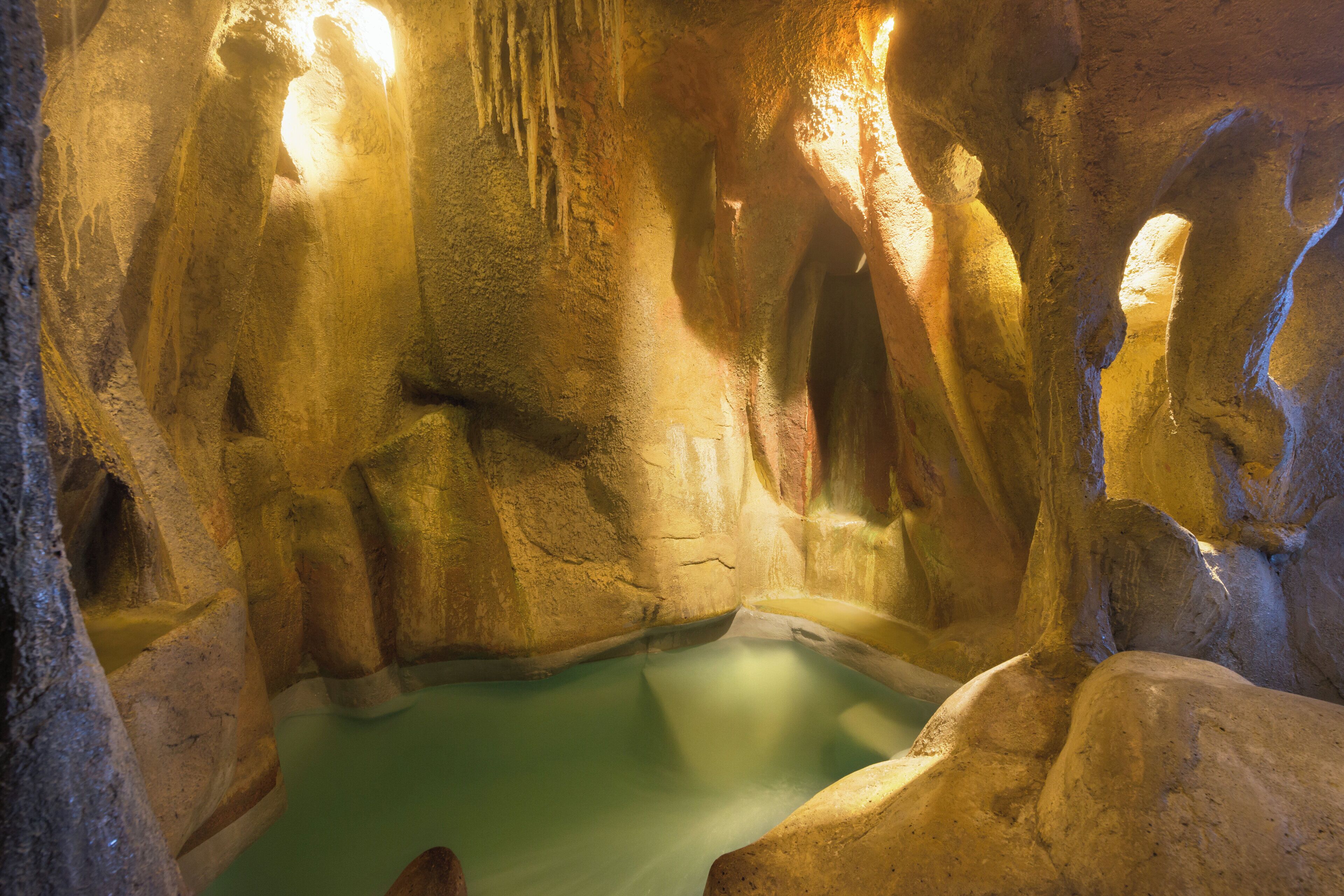A cave with stalactites and stalagmites, a pool of water, and warm lighting.
