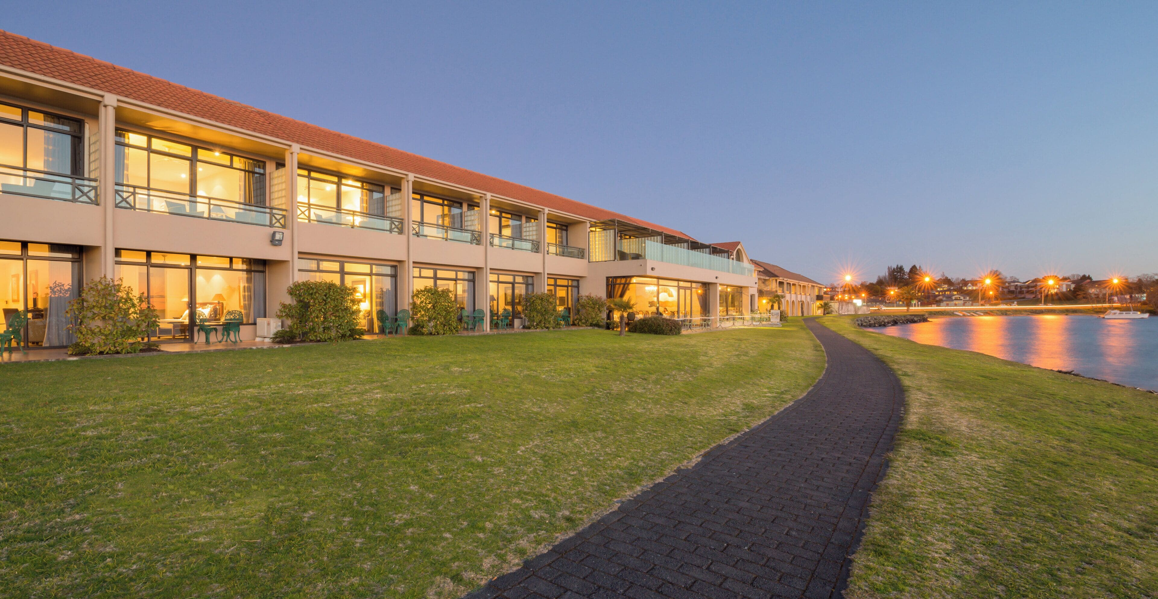 A hotel with a paved walkway leading to a body of water, multiple balconies, and outdoor seating areas.
