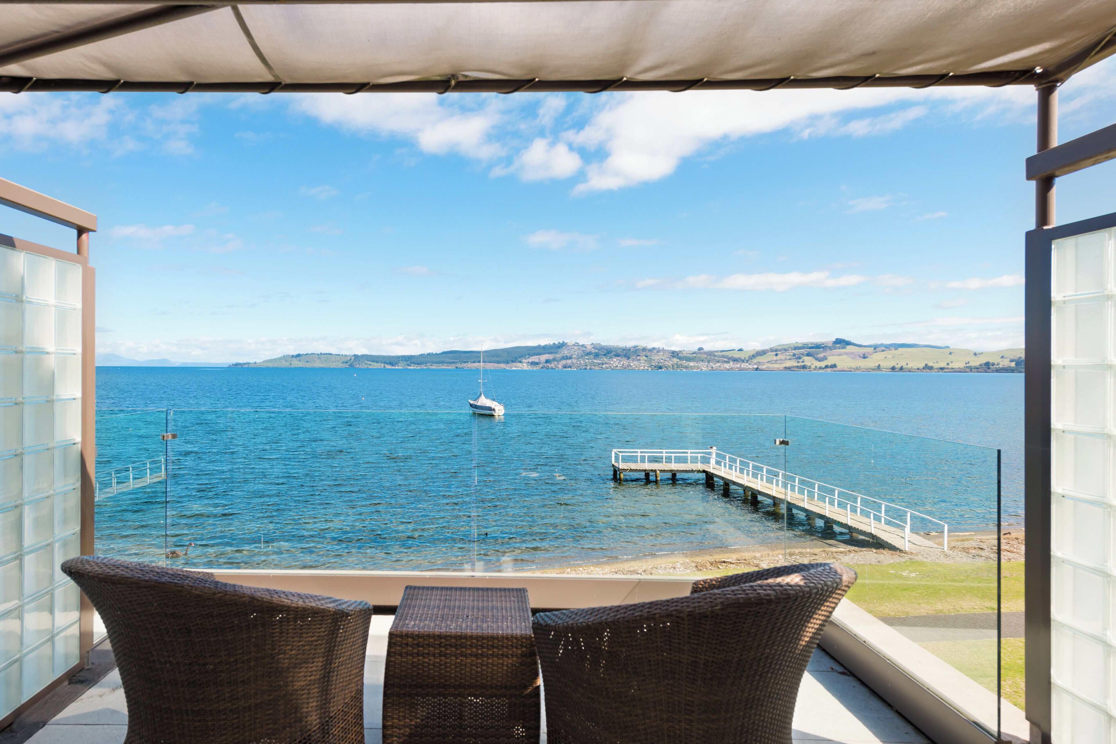 A balcony with wicker furniture overlooking a body of water, a dock, and distant hills.